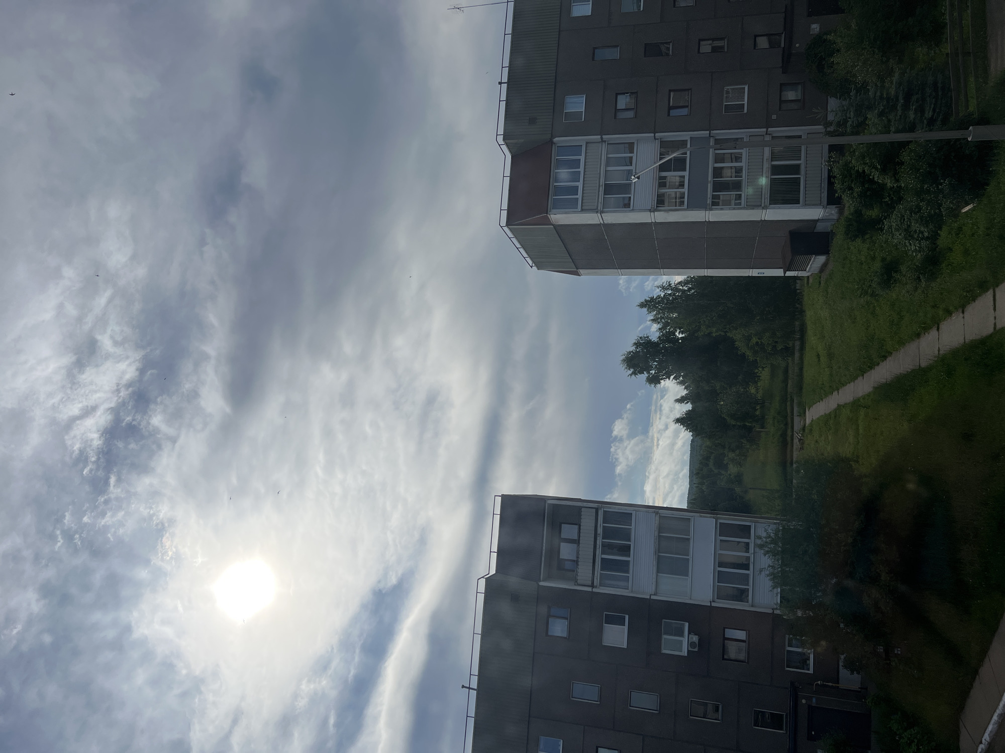 The image captures a view of two residential apartment buildings in the city of Марковский, Russia, framed by a sky with scattered clouds and a bright sun. The buildings are multi-story, with dark gray facades and numerous windows. The one on the left appears to have a balcony or an extended section with white frames. The building on the right has a pitched roof with a reddish-brown section and a balcony with white window frames.
Between the buildings, a grassy area slopes upwards, dotted with trees. A concrete pathway runs through the grass, leading towards the distant trees and hills. The overall impression is that of a residential area with surrounding natural landscape. There are no people visible in the image, and the environment appears to be calm and uneventful. The lighting suggests it is daytime, possibly late morning or early afternoon given the sun's position.