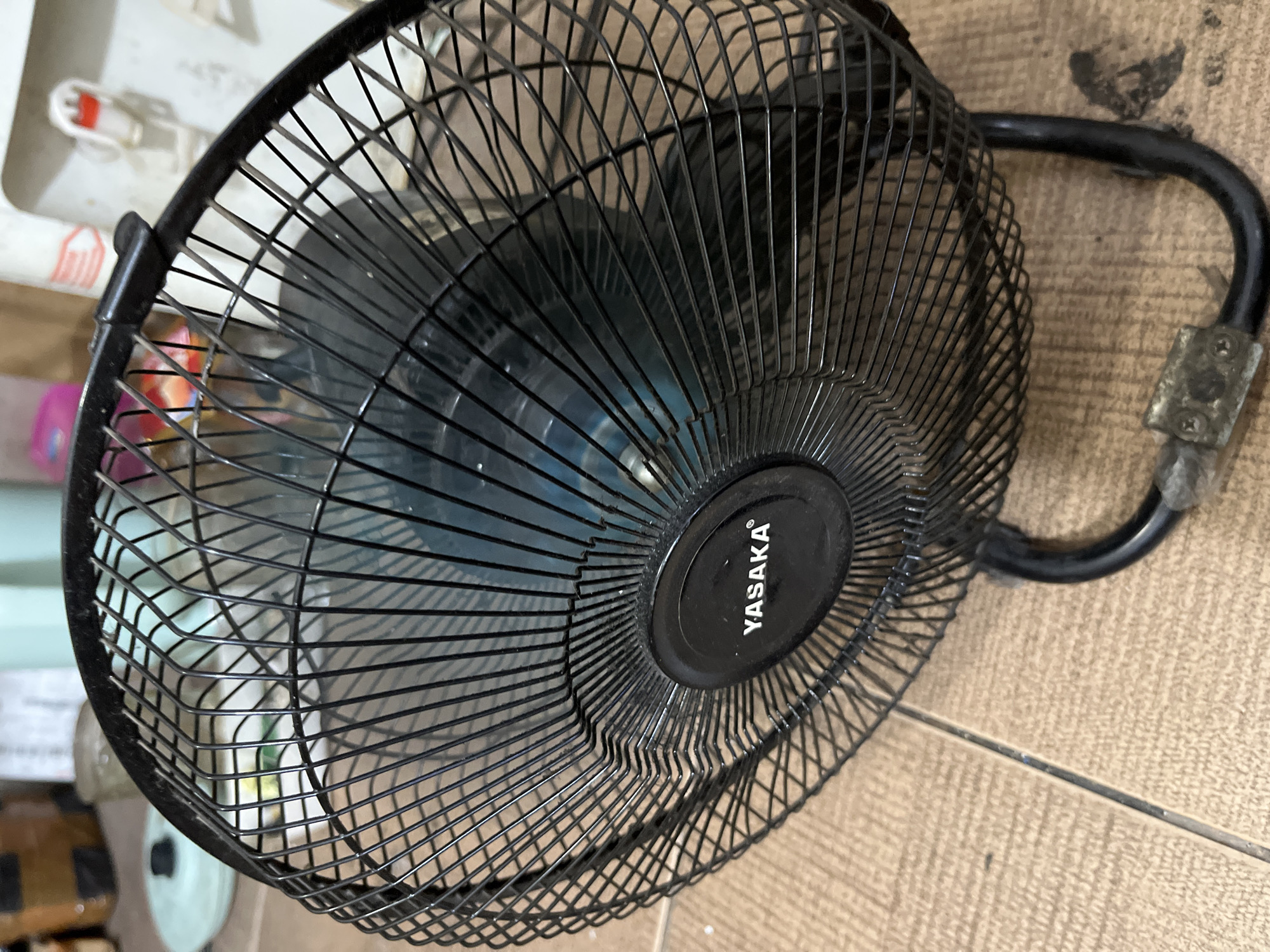 The image shows a black oscillating fan with the brand name "YASAKA" visible on its circular housing. The fan is positioned on a tiled floor with a brown, wood-grain pattern. In the background, out of focus, are various household items including a water dispenser, boxes, and a light blue lid. The scene appears to be indoors, likely in a residential setting within Central Jakarta, Indonesia, judging by the typical flooring and common household appliances seen. There are no people or discernible actions taking place. The lighting is ambient, suggesting an indoor environment.