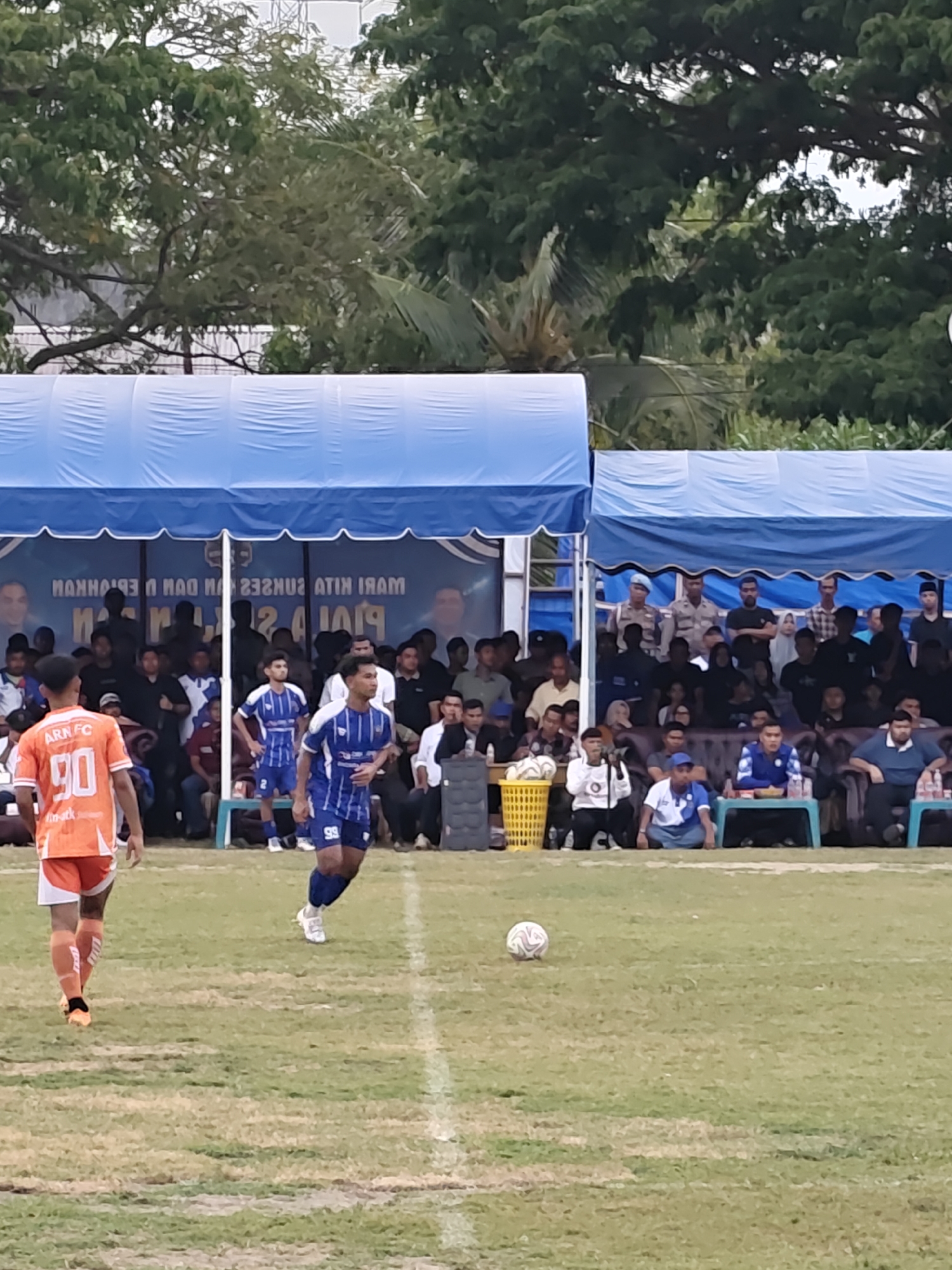 The image depicts an outdoor soccer match taking place on a grass field in Desa Meunasah Blang, Indonesia. The foreground shows two soccer players. The player on the left is wearing an orange jersey with "ARN FC 90" printed on the back and orange shorts with white trim. The player on the right is wearing a blue and white striped jersey with the number "99" on the shorts and blue shorts. A soccer ball is visible on the grass between the players. In the background, spectators are seated under blue tents. The tents have white trim and are supported by poles. A banner is visible behind the spectators with text in what appears to be Indonesian. Trees are visible behind the tents, suggesting a location with natural surroundings. The overall scene is characteristic of a local sporting event.