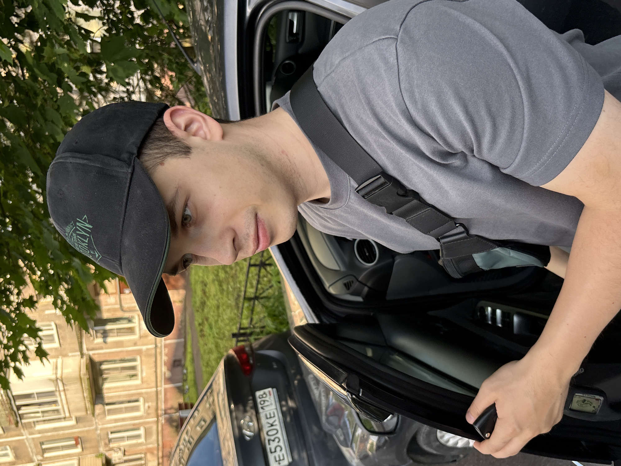 The image shows a young Caucasian male wearing a black baseball cap and a grey t-shirt, standing next to an open car door in what appears to be a residential area. The man is holding a car key in his right hand. A dark-colored car is parked to his left. In the background, a multi-story apartment building with light-colored facades is visible, along with green trees and a street. The presence of a Hyundai car with Russian license plates, particularly the "E530KE198" plate, strongly suggests the location is in Saint Petersburg, Russia, as the "198" region code corresponds to the Leningrad Oblast, which includes Saint Petersburg. The overall scene depicts a common urban setting on what appears to be a sunny day.