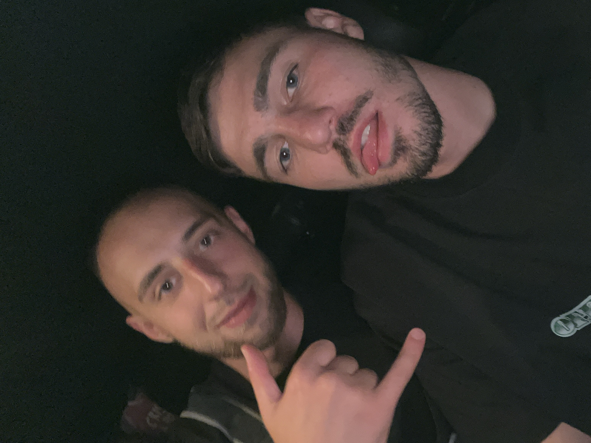 Two young men are in the foreground of a dimly lit image, possibly taken inside a vehicle. The man on the right has light brown hair, blue eyes, a beard, and is sticking his tongue out. He is wearing a black t-shirt. The man on the left has a shaved head and is smiling. He is also wearing a black t-shirt and making a "rock on" hand gesture. The background is dark and out of focus.
