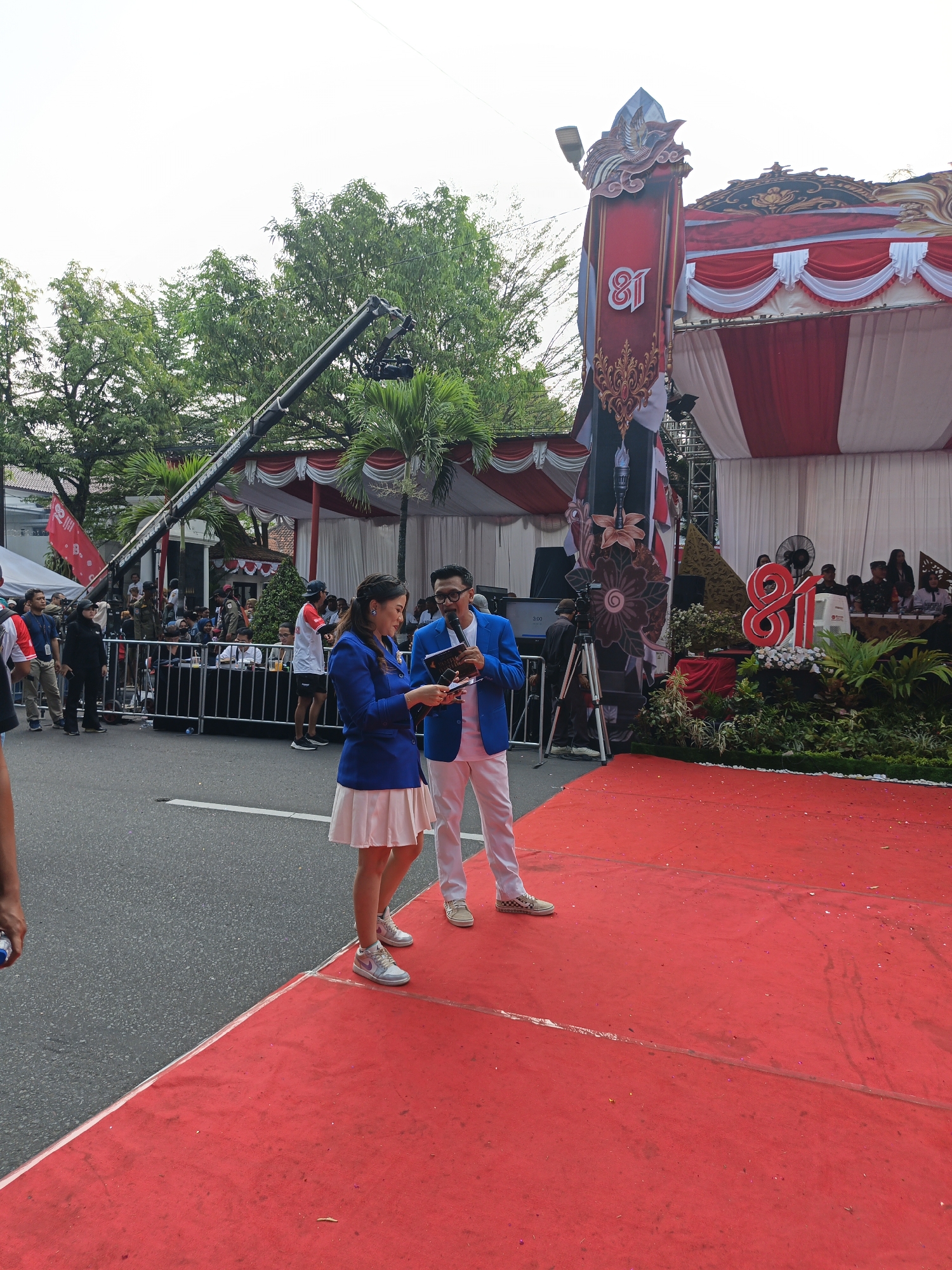 In the city of Klaten, Indonesia, a red carpet event is underway, possibly a celebration or broadcast. Two individuals, a woman in a blue blazer and white skirt and a man in a blue blazer and white pants, stand on the carpet, appearing to engage in a presentation or interview. The woman holds a booklet, and the man holds a microphone and a similar booklet. Behind them, a large stage is decorated with red and white drapery and a prominent "81" numeral, suggesting an anniversary or significant milestone. Trees and a cloudy sky form the backdrop. A camera crane is positioned overhead, indicating media presence. A crowd of onlookers and event staff are visible behind a metal barricade.