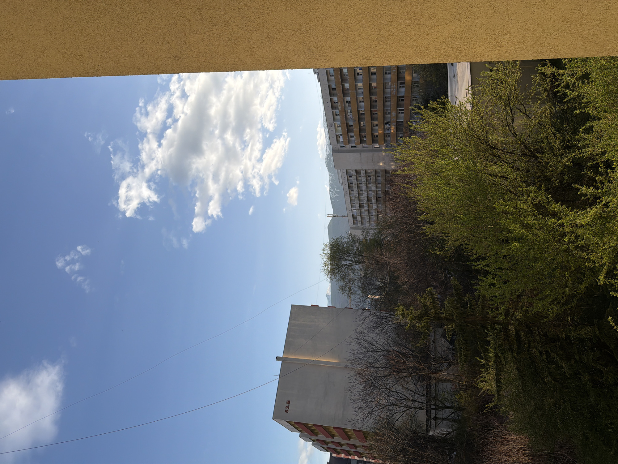 A ground-level, outdoor view of a cityscape in Sofia, Bulgaria, framed by a yellow-painted balcony ceiling. The sky is blue with scattered white clouds. Buildings of varying heights are visible, including a taller, beige-colored building with numerous windows. Trees with green foliage are on the right. A light-colored building with the number "53.5" in red is on the left, below, with power lines running across the sky. Mountains are faintly visible in the distance, between buildings.