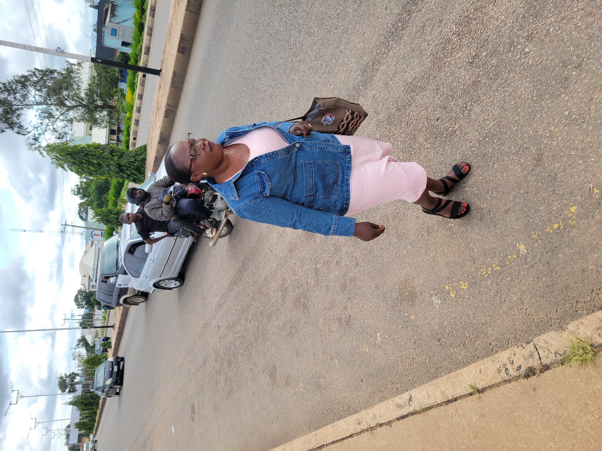 A woman walks on an asphalt road in Jos, Nigeria, with a cityscape in the background. She is wearing a blue denim jacket over a pink dress and black sandals. She carries a brown tote bag in her right hand. Behind her, a motorcycle with two passengers drives away. Further down the road, a silver car and a black SUV are visible. The environment appears to be a moderately busy street with scattered trees and buildings. The sky is overcast.
