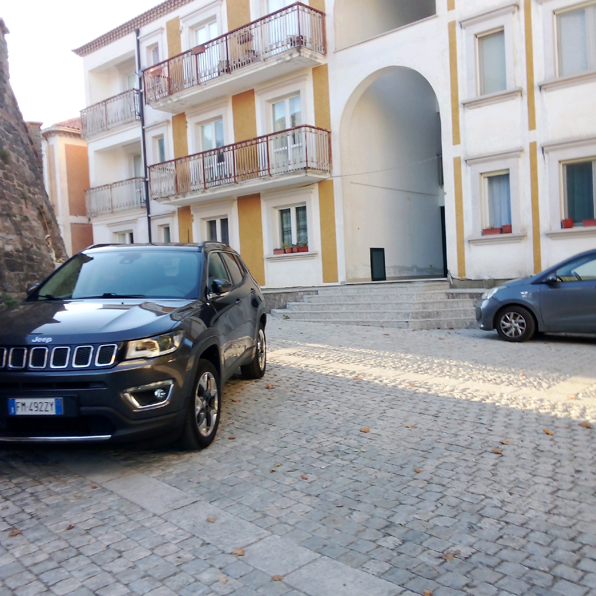 A dark grey Jeep Compass is parked on a cobblestone street in front of a multi-story building. The building has white and yellow facades with balconies. A darker grey car is parked further down the street. The image appears to be taken during the daytime. The location is identified as Castelnuovo di Conza, Italy.