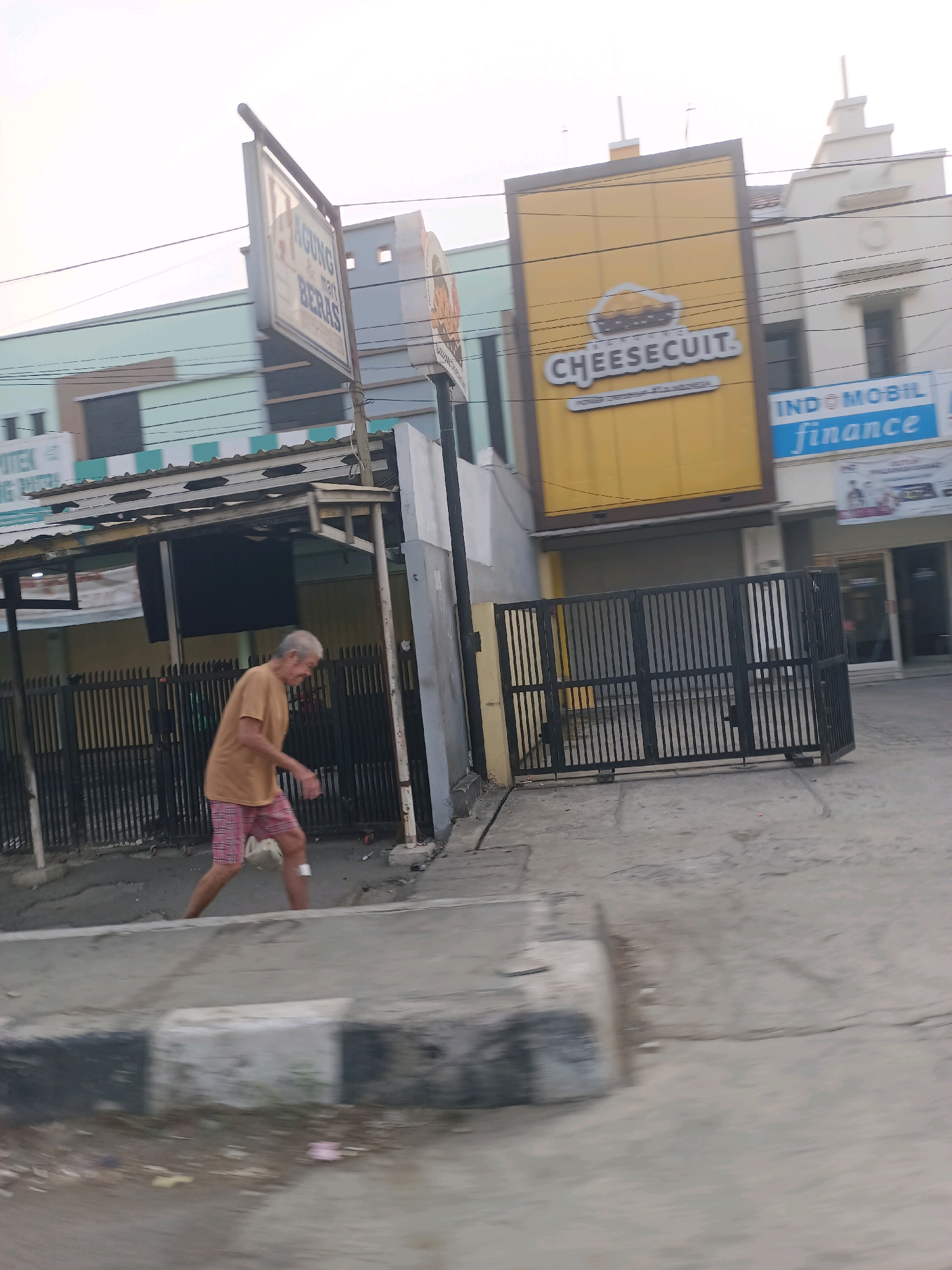 In Karawang, Indonesia, an elderly man with graying hair walks past a gated property. He is wearing a light brown t-shirt and plaid shorts. A white bag hangs from his left leg. The property features a building with a large yellow sign that reads "CHEESECUIT." Next to it, a building displays a blue sign for "INDO MOBIL finance." A street sign with red lettering is also visible. The area appears to be a commercial district with various businesses.