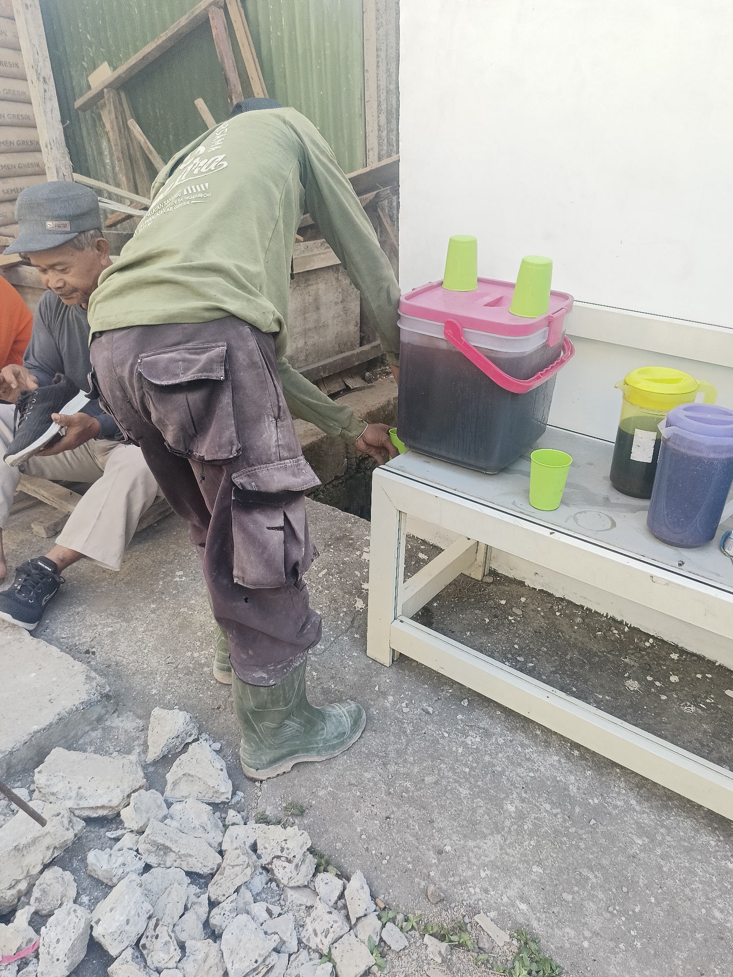 The image depicts an outdoor scene in Gedangan, Indonesia, likely near a construction or repair site, given the debris of broken concrete in the foreground. Two men are present. One man, wearing a green long-sleeved shirt, brown cargo pants, and green rubber boots, is bending over, appearing to interact with something on the ground near a white framed structure. The other man, to the left, wears a grey t-shirt, light-colored pants, and a dark baseball cap. He is seated and holding a black shoe.
On the white framed structure, which resembles a table or bench, are several containers holding beverages. A large pink-lidded container holds a dark liquid and has two green cups resting on its lid. To its right, a yellow-lidded pitcher and a purple-lidded pitcher also contain dark liquid, with a single green cup placed in front of them. The background shows construction materials, including stacked bags labeled "SEMEN GRESİK," a common cement brand in Indonesia, and some wooden framing, suggesting ongoing work. The ground is paved with concrete. The lighting suggests it is daytime.