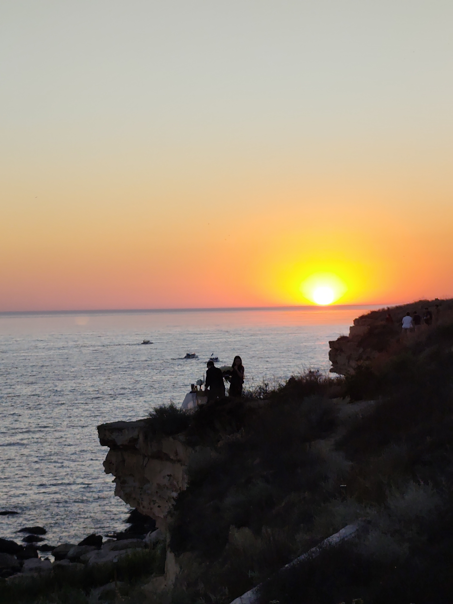 The image depicts a sunset over the Caspian Sea near Aktau, Kazakhstan. The sky transitions from a pale yellow-white at the top to vibrant oranges and yellows as it nears the horizon, where the sun, a bright yellow orb, is setting. The sea below is a deep blue with gentle waves.
Along the rugged, rocky coastline, several small boats are visible on the water. On the cliff edge in the foreground, silhouetted figures are gathered. On the left side of the cliff, two figures are standing near what appears to be a table set for a meal, suggesting a special occasion or a romantic dinner. Further to the right, on the same cliff, a group of people are standing and observing the sunset. The vegetation on the cliff is dark and appears bushy. The overall scene is one of natural beauty and human enjoyment of the twilight hours.