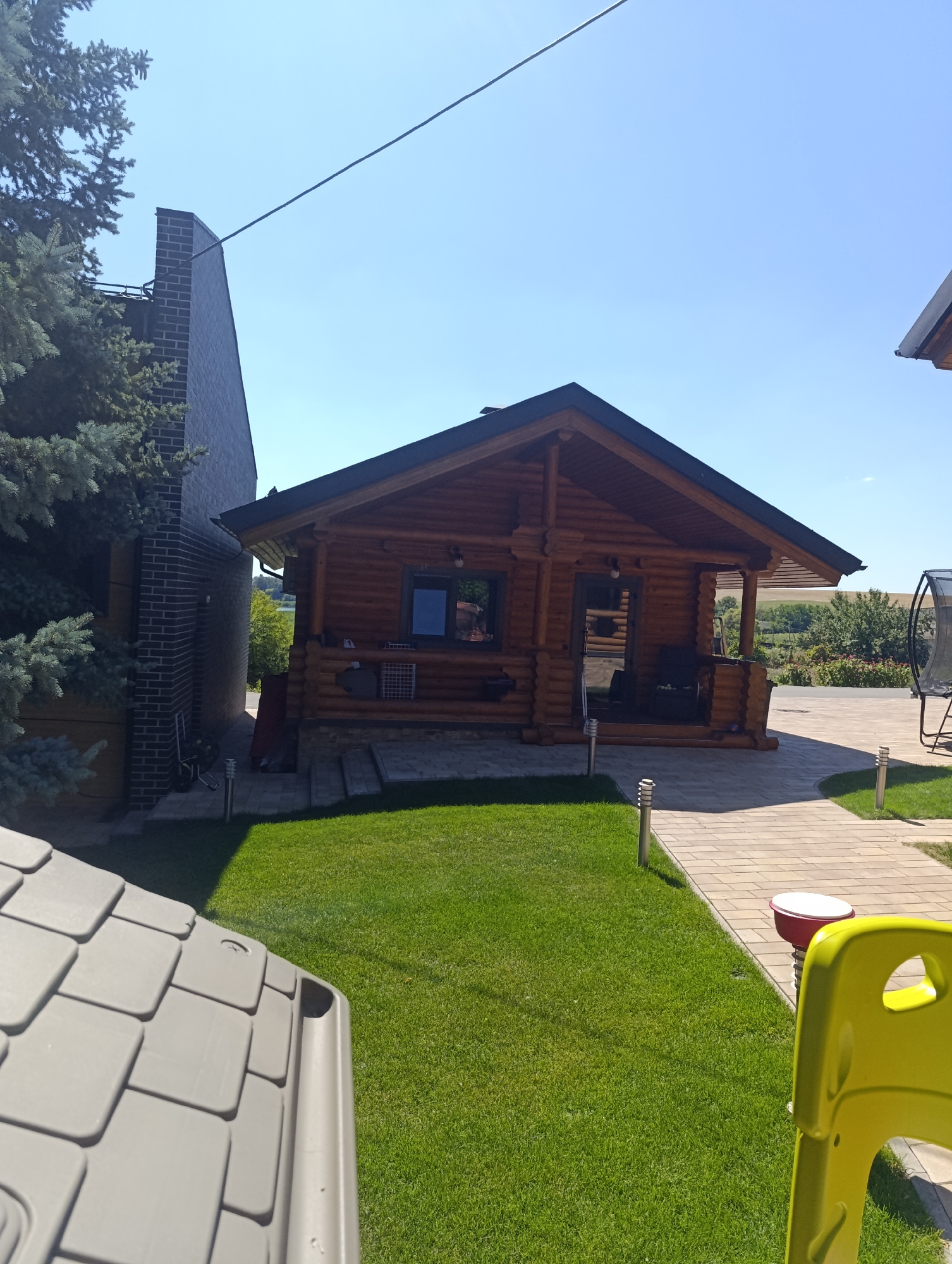 In the city of Bulatselivka, Ukraine, a scene unfolds under a clear blue sky. A wooden log cabin with a dark roof stands prominently, featuring a porch with seating. To the left of the cabin, a tall, dark brick structure, possibly a chimney or an extension, rises high. In the foreground, a section of a grey tiled roof, likely from a smaller structure, is visible on the left. A neatly manicured green lawn stretches out in front of the cabin, bordered by a pathway made of light-colored interlocking pavers. Several modern, minimalist light fixtures are placed along the pathway. To the right, a bright yellow plastic chair and a red and white object, possibly a trash receptacle, are situated near the path. In the background, lush greenery and trees suggest a rural or suburban setting. The overall impression is one of a well-maintained outdoor living space.