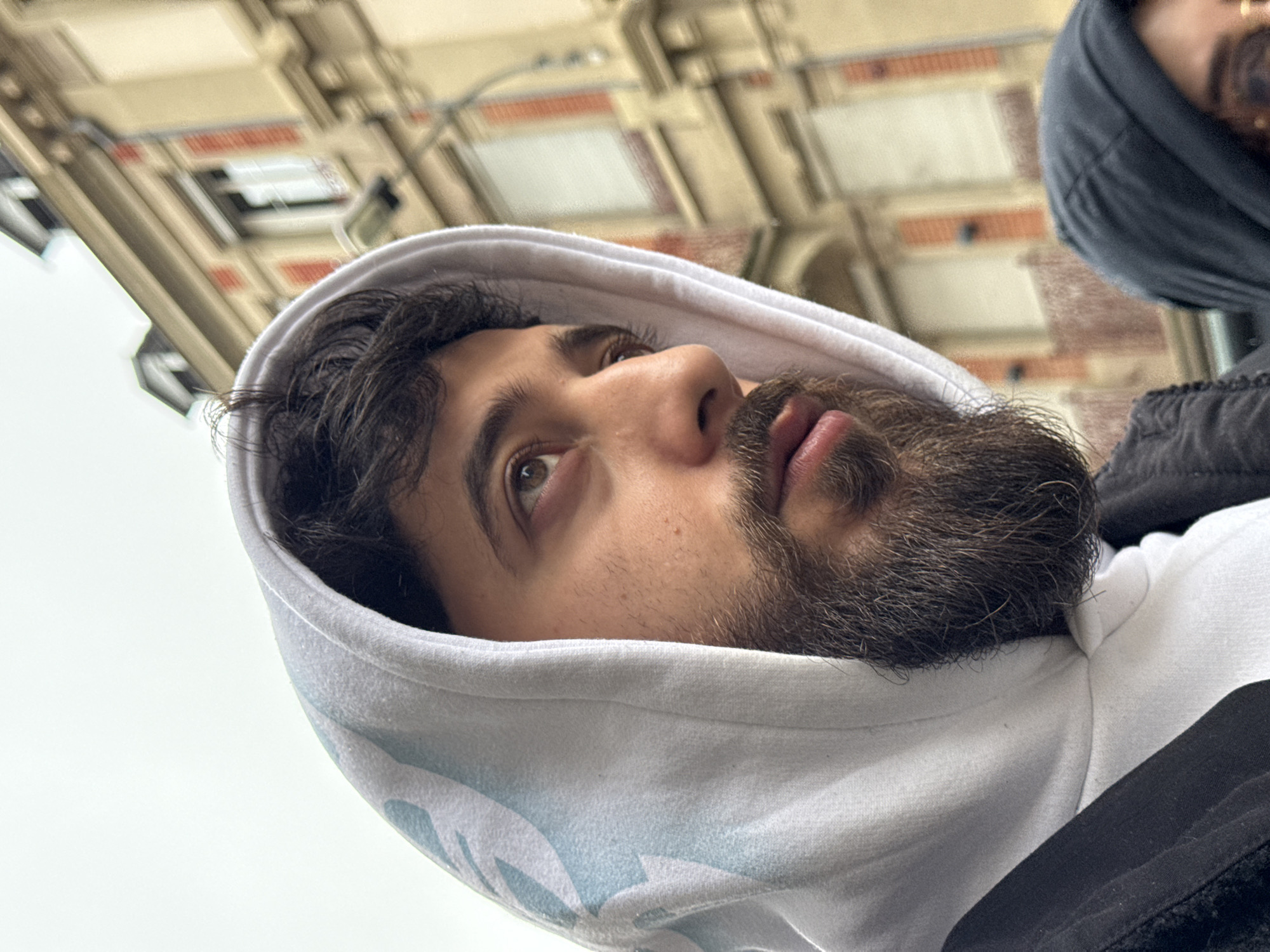 The image displays a close-up perspective of an adult male in an urban outdoor setting. The man, positioned prominently in the foreground, has dark hair, a full dark beard, and brown eyes, which are directed upward and to the viewer's left. He is wearing a white hooded sweatshirt with a faint light-blue pattern, layered under a dark-colored jacket. A second individual is partially visible in the lower right corner, obscured by the main subject, wearing a dark hooded garment. The background consists of a multi-story building characterized by a light-colored, possibly cream or beige, facade adorned with vertical red brick accents. Visible elements of the building include dark-framed windows, some with rolled-down shutters, and a utility pipe or downspout. The sky above is overcast, appearing light grey. The architectural style, with its combination of light facades and red brick detailing, is typical of structures found in the city of Strasbourg, France.