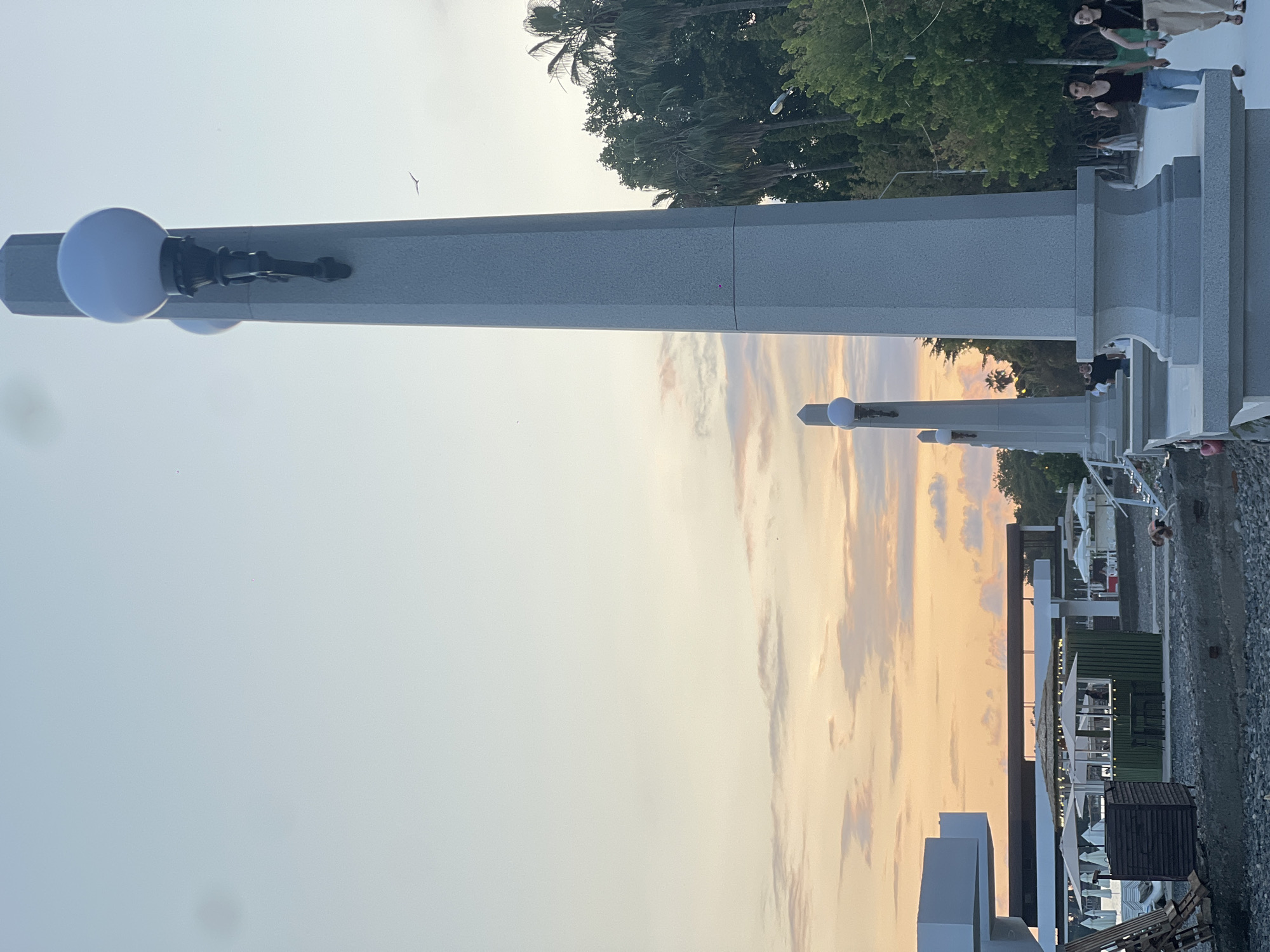 This photograph, taken in Sokhumi, Abkhazia, depicts a picturesque scene at dusk along a waterfront promenade. The foreground is dominated by a large, grey, polygonal lamppost with a white spherical lamp. To the left of the lamppost, a row of similar lampposts recedes into the distance. Beyond the lampposts, there are several structures, including a modern white building with geometric lines and a green kiosk with outdoor seating. Umbrellas and lounge chairs are visible on what appears to be a pebbly beach. In the middle ground, a paved walkway is populated by a few individuals, including two young women in casual attire walking together. The background features a dense line of palm trees and other lush green foliage, suggesting a tropical or subtropical climate. The sky above is a soft gradient of pale blue and peach, with wispy clouds indicating the setting sun. A small bird is captured in flight against the vast expanse of the sky. The overall atmosphere is tranquil and serene, characteristic of a peaceful evening by the sea.