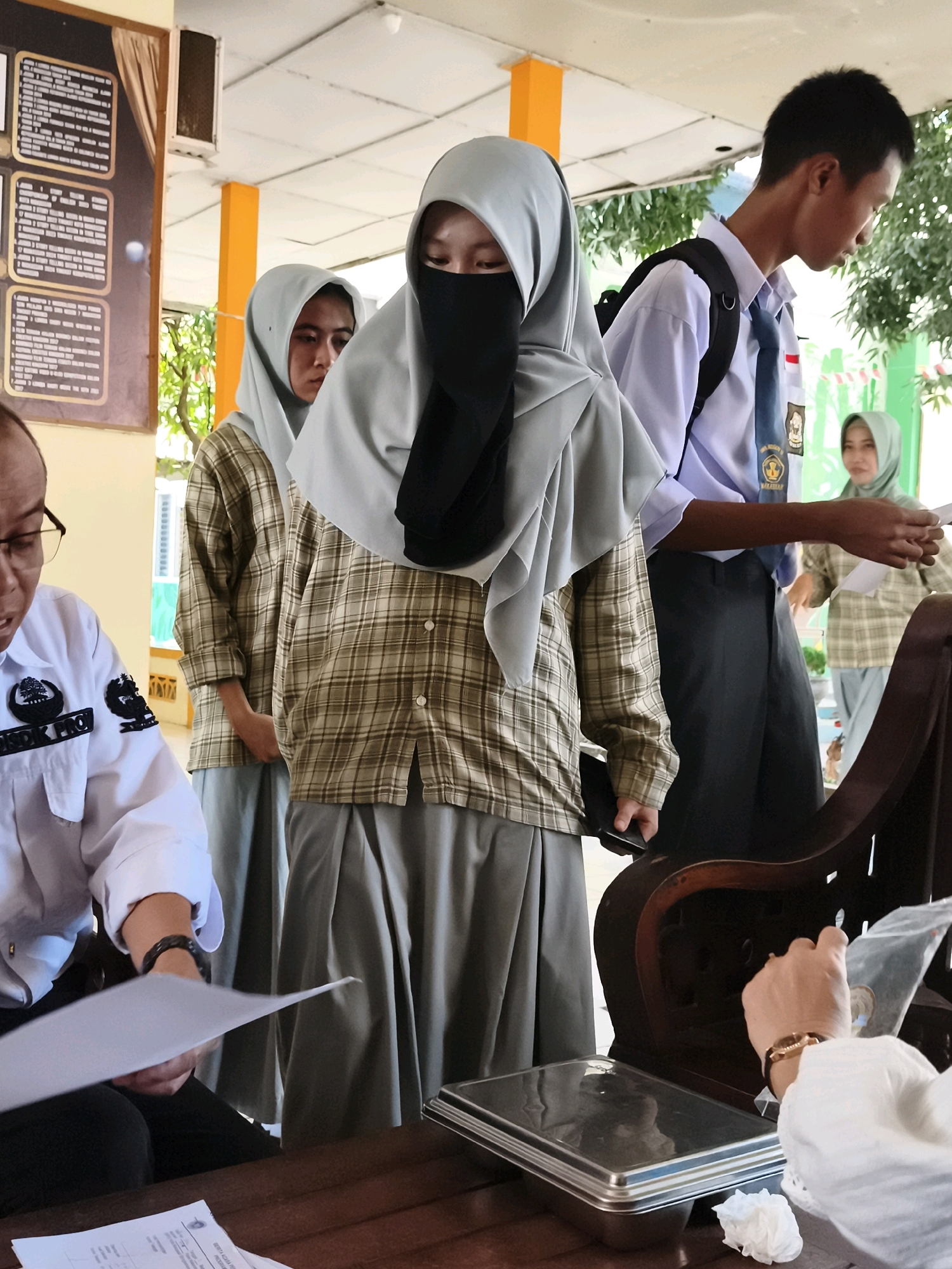 The image depicts a scene at a school in Makassar, Indonesia. In the foreground, a person in a white shirt with a name tag that reads "DISDIK PROV" is seated at a wooden table, looking down at papers. They are holding up a piece of paper. To the right, a hand with a bracelet is reaching into a plastic bag, and next to it is a crumpled white tissue. On the table, there's a metallic container and more papers. Behind the person in the white shirt, several students are standing. Two female students in headscarves and plaid shirts are visible, with one wearing a black niqab. A male student in a school uniform, including a blue tie, stands to the right, with a backpack on. In the background, another female student in a headscarf and plaid shirt is partially visible. The environment suggests an outdoor covered area, possibly a school hallway or entrance, with a bulletin board on the left and greenery visible through openings in the structure.