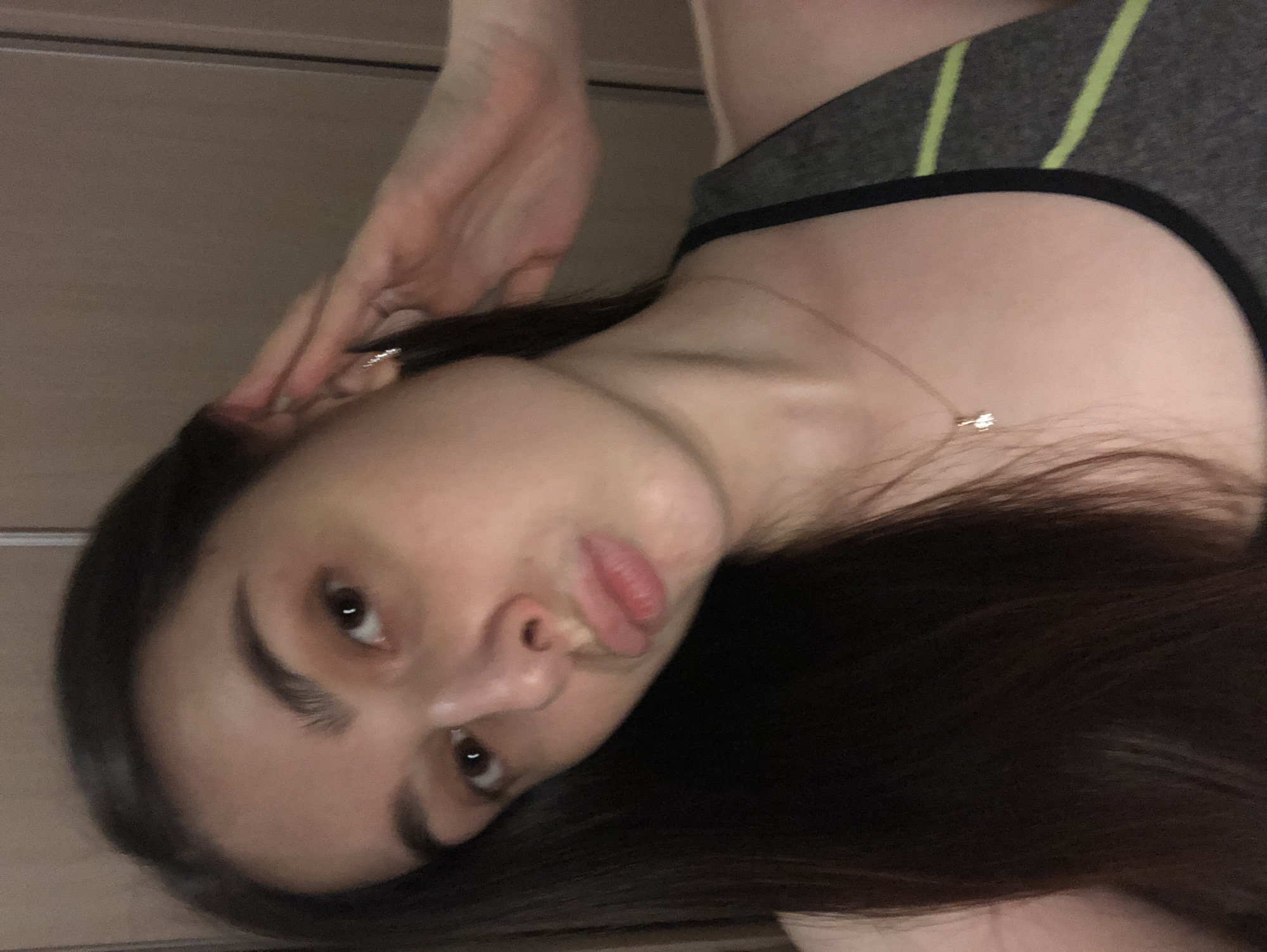A young woman with dark hair is positioned closely to the camera. She is wearing a gray top with black trim and light green stripes near the top of the shoulders. She is also wearing a delicate necklace. Her hand is raised near her face. The background consists of what appears to be a wooden wall. The image location is unknown.