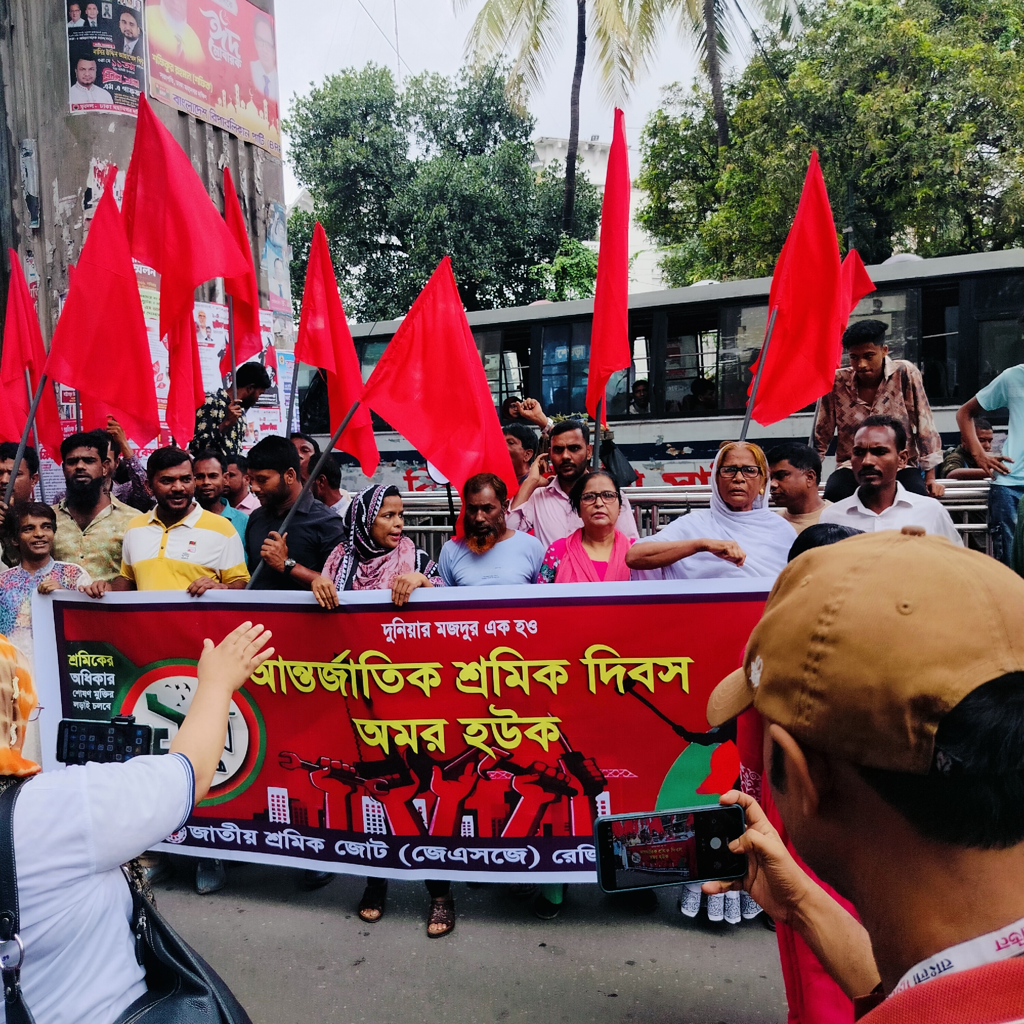 A street-level photograph taken in an urban setting, potentially in the city of Unknown, Unknown. The scene depicts a procession or demonstration, with a large group of people marching and holding red flags, which are prominent throughout the frame. A large banner, predominantly red, is held at the front of the crowd. The banner includes text in a language that appears to be Bengali. The text translates to "Long live international workers' day" and the banner bears the name of the organization "Jatio Sramik Jote". The environment includes buildings with posters and advertisements. A bus is visible in the background, with people standing on its platform. In the foreground, an individual on the right wearing a cap and a red shirt is holding a smartphone and taking a photo of the marchers. On the left side of the foreground, another individual holds a smartphone, also seemingly recording the event. The atmosphere suggests a political or labor rights rally. The weather appears to be overcast or slightly hazy.