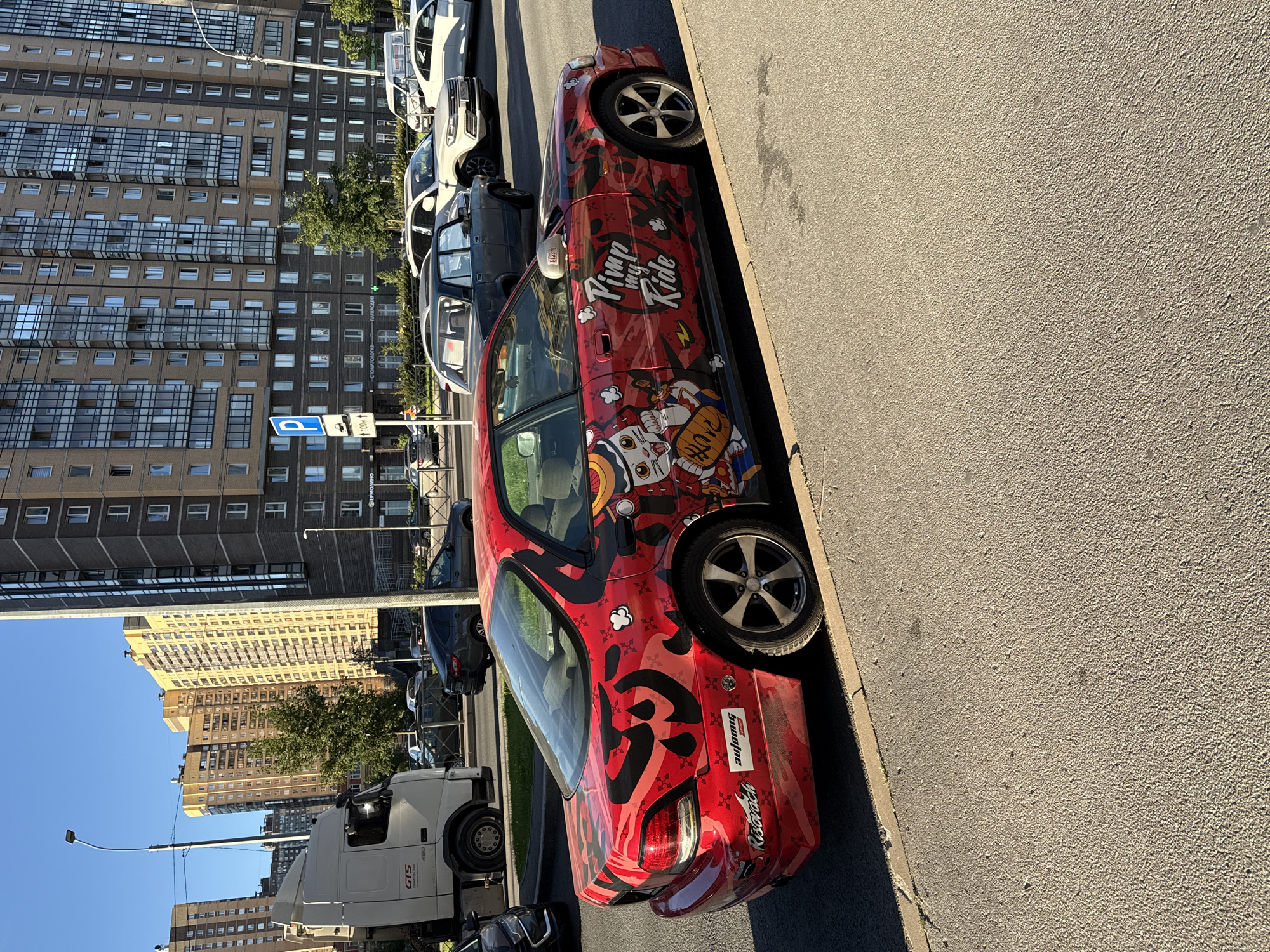 A red sedan with anime-style graphics, including a cat character and the text "Pimp my Ride", is parked on the side of a street in Pargolovo, Russia. The car has custom wheels and stickers on its rear bumper. In the background, there are several tall apartment buildings, a white truck, and other vehicles on the road. The sky is clear and blue, and the sun is casting shadows. The image appears to be taken during daylight hours.