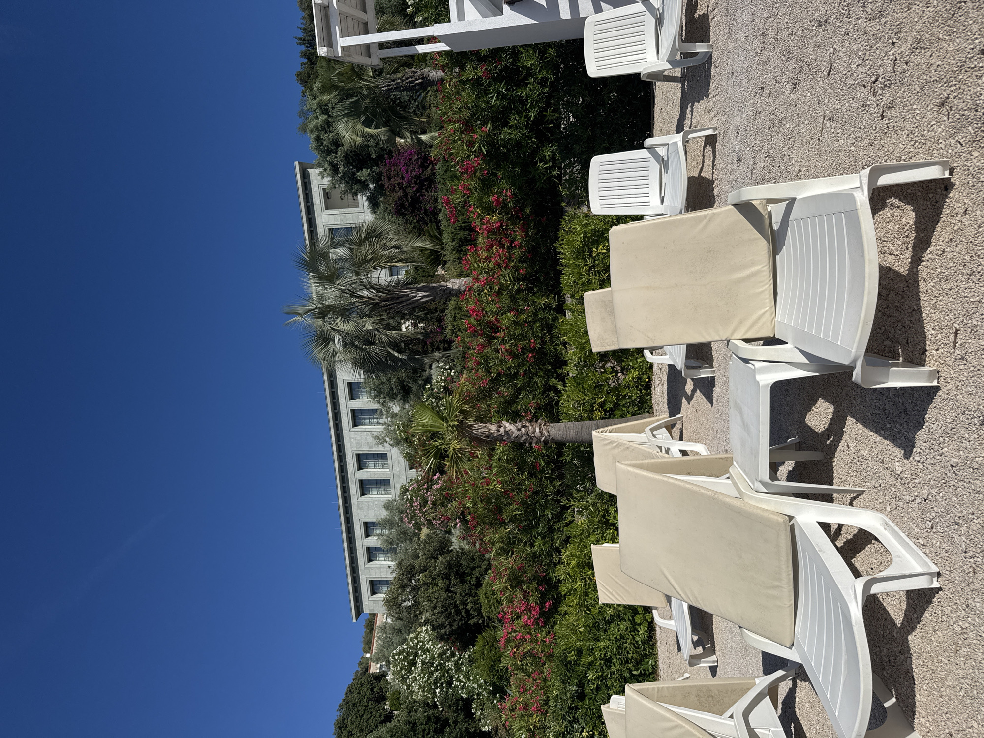 The image depicts an unoccupied outdoor leisure area on a clear, sunny day in the city of Unknown, Unknown. The scene is dominated by a clear, deep azure sky that occupies the upper portion of the frame. Below the sky, a multi-level, light grey stone-clad building with numerous rectangular windows with dark frames is situated atop a densely vegetated hillside. The hillside features a rich array of green foliage, including various broadleaf trees, slender palm trees, and flowering shrubs with clusters of vibrant pink, red, and white blossoms, creating a lush, tropical appearance. To the far right, a section of a white wooden pergola or structure is partially visible.
In the foreground, a ground surface composed of light-colored, fine gravel or small pebbles is visible. Several white plastic reclining lounge chairs are arranged on this gravel, each fitted with a removable, light cream-colored padded cushion on the seat and backrest. These chairs are empty and positioned in a manner suggesting preparation for occupancy. A small, white plastic side table is placed between two chairs in the lower center of the frame. The bright sun casts distinct shadows from the chairs and table onto the gravel. No people or animals are present in the image, and no active interactions or specific actions are occurring. The overall environment suggests a tranquil resort or private beach setting.
