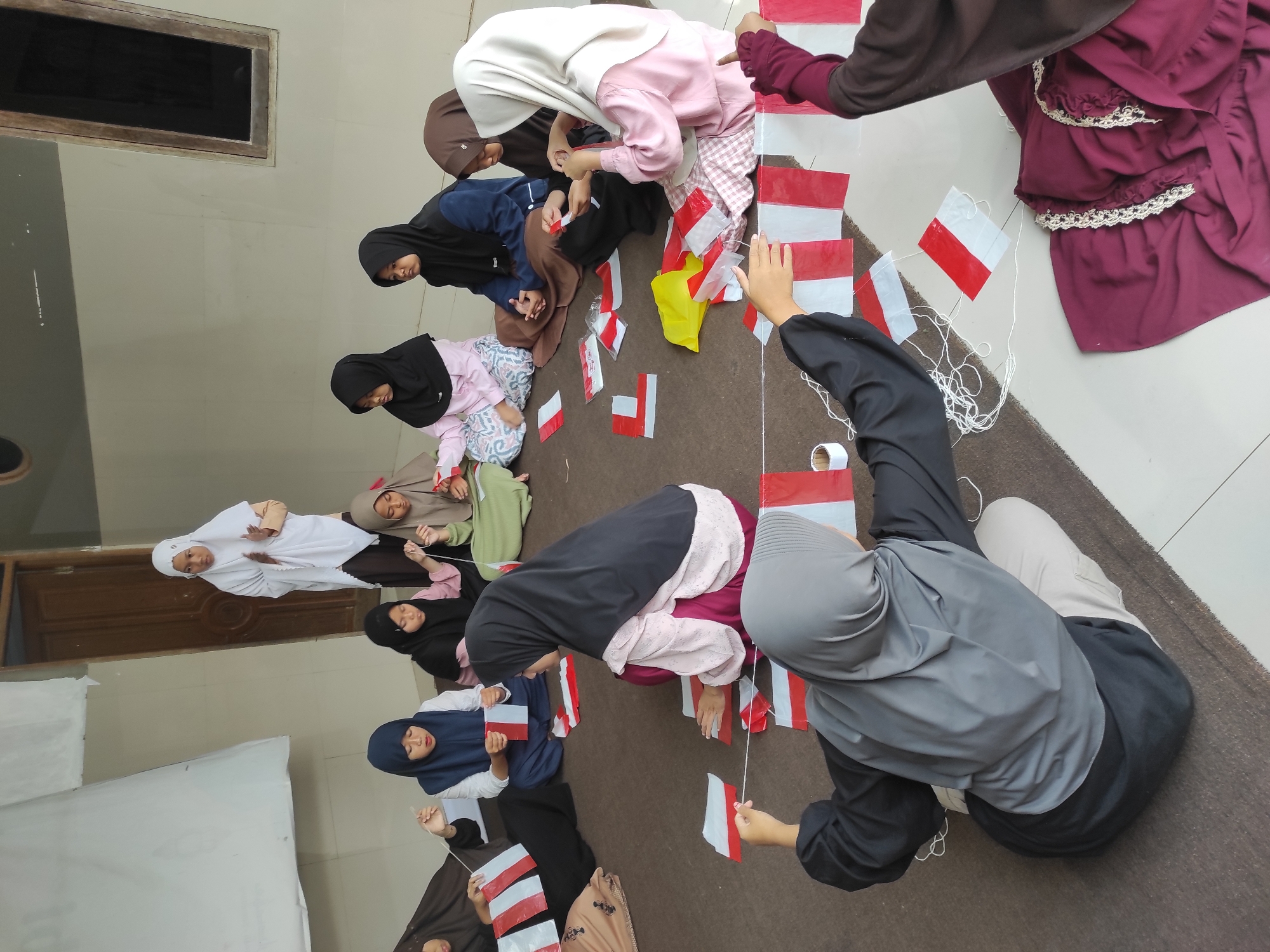 In Sumedang, Indonesia, a group of approximately ten young women and girls are gathered indoors, sitting on the floor and engaged in a crafting activity. They are in the process of making small red and white flags, likely for a national celebration given the colors. Most of the participants are wearing hijabs in various colors, including white, black, blue, brown, and gray. Several individuals are holding pieces of red and white fabric or paper and string, with the flags strung together in a line. A woman standing in the background, also wearing a white hijab, appears to be supervising or instructing the group. The environment suggests a communal indoor space, possibly a community hall or a large room, with a textured brown floor and a tiled wall in the background. The lighting is natural, indicating the photo was taken during the daytime.