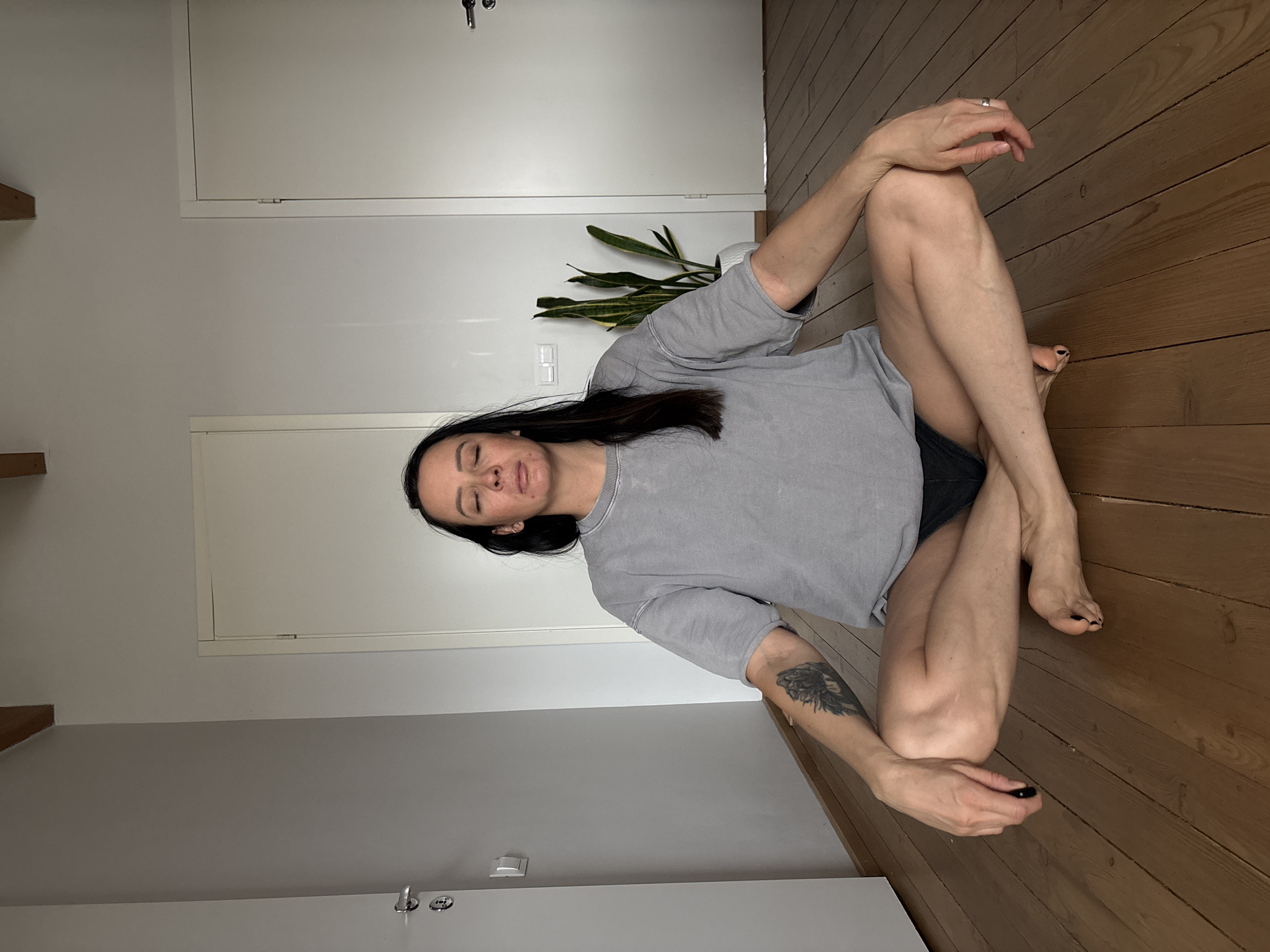 A woman sits in a cross-legged position on a wooden floor in an indoor setting. She wears a grey t-shirt and dark shorts. Her hair is dark and straight. She has a tattoo on her left arm. The room has white walls and includes two doors. A plant is positioned between the doors. The location is not identifiable as the city of Chaikovsky, Russia from visual information provided.