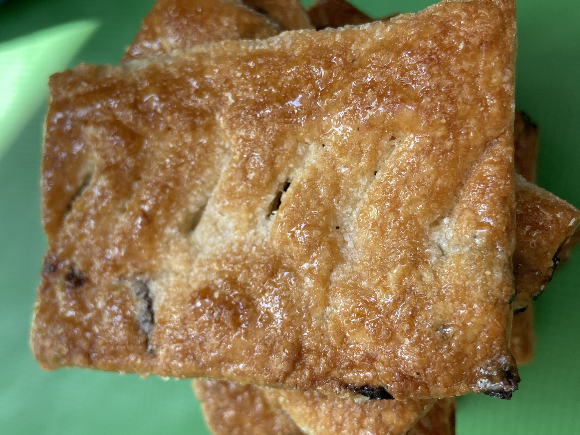 A close-up, overhead shot shows a stack of golden-brown, square-shaped pastries on a bright green background. The pastries have a flaky, slightly greasy appearance with visible sugar crystals and some darker, possibly fruit-filled, inclusions peeking through the cut-out slits on the top surface. The lighting casts subtle shadows and highlights, emphasizing the textured surface of the baked goods. The background is a solid, vibrant green, out of focus. There are no people or any other discernible objects in the scene.