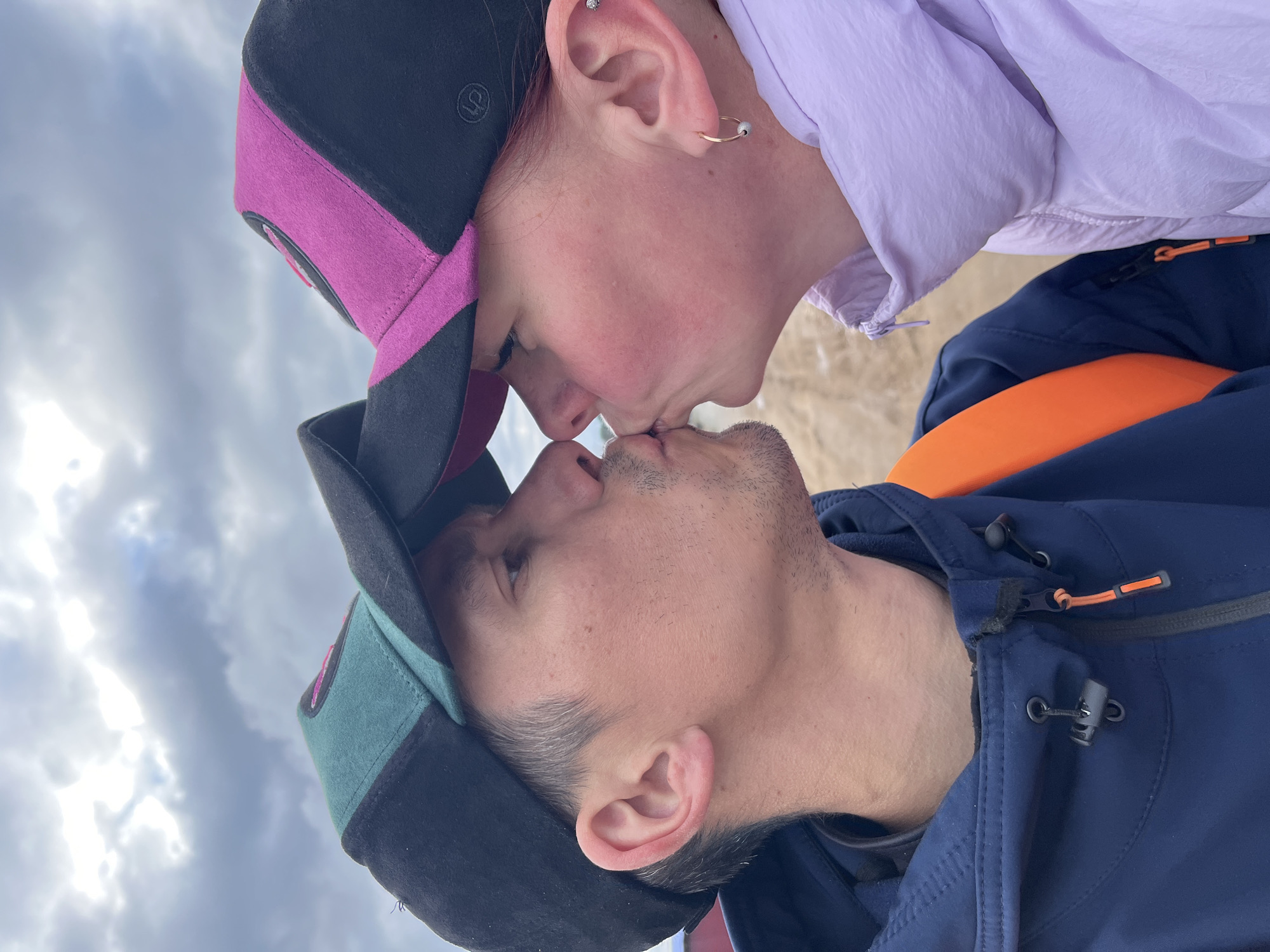 The image captures a close-up, intimate moment between two individuals sharing a kiss outdoors, photographed from a low angle, looking upwards towards them against a cloudy sky.
One individual, positioned at the top of the embrace, wears a black baseball cap with magenta brim and side panels. An embroidered stylized 'U' or Omega symbol is visible on the cap. This person also wears a light purple jacket and has several ear piercings, including a silver stud in the helix and a gold hoop in the lobe. Their face appears slightly flushed.
The other individual, positioned at the bottom, wears a black baseball cap with teal or dark green brim and side panels. They have short facial hair and are dressed in a navy blue jacket with visible orange accents, such as a zipper pull and an internal orange lining or strap.
Their lips are pressed together in a tender kiss, conveying affection and intimacy. The background predominantly features a cloudy, bright sky, indicating an overcast daytime setting. A hint of light brown or beige ground, possibly sand or earth, is visible behind the upper individual. The jackets worn by both subjects suggest cooler weather conditions. The overall scene is consistent with an outdoor activity in a location like Chaikovsky, Russia, perhaps near a body of water or open landscape.