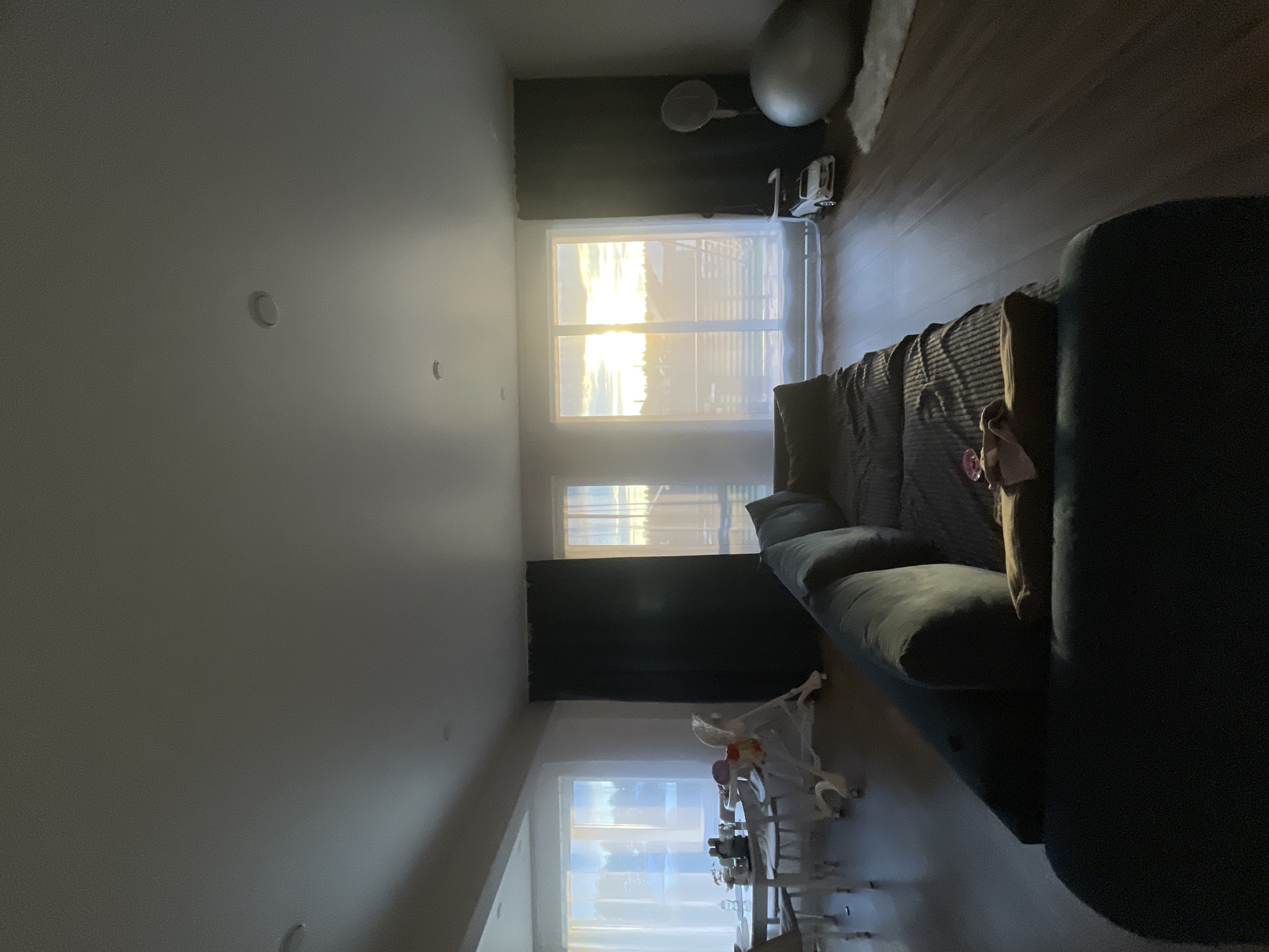 This media file captures an indoor scene, possibly a living room, with a strong emphasis on natural light entering through windows. The dominant objects are a large, dark upholstered sofa that occupies a significant portion of the frame, and a dining set with chairs and a high chair, suggesting a family or domestic setting.
The scene is illuminated by the warm, golden light of what appears to be a sunset, indicating the time of day. The light streams through two large windows, casting long shadows and creating a bright, almost ethereal atmosphere. One window shows a hazy view of what could be a landscape with a body of water and some structures. The other window, positioned lower, also lets in a significant amount of daylight.
Other visible objects include a large, grey exercise ball near one of the windows, and a white and grey object that appears to be a modern lamp or light fixture. A dark curtain hangs by the window on the left, contrasting with the bright light. There are some small, circular indentations on the ceiling, possibly recessed lighting fixtures.
There are no people visibly present in the frame, but the presence of the high chair implies the potential for a child to be in the room. The overall mood is serene and peaceful, largely due to the quality of the light. The location cues, combined with the provided context, suggest this might be a home in Podshivalovo, Russia, during sunset. No visible text is present in the image.
