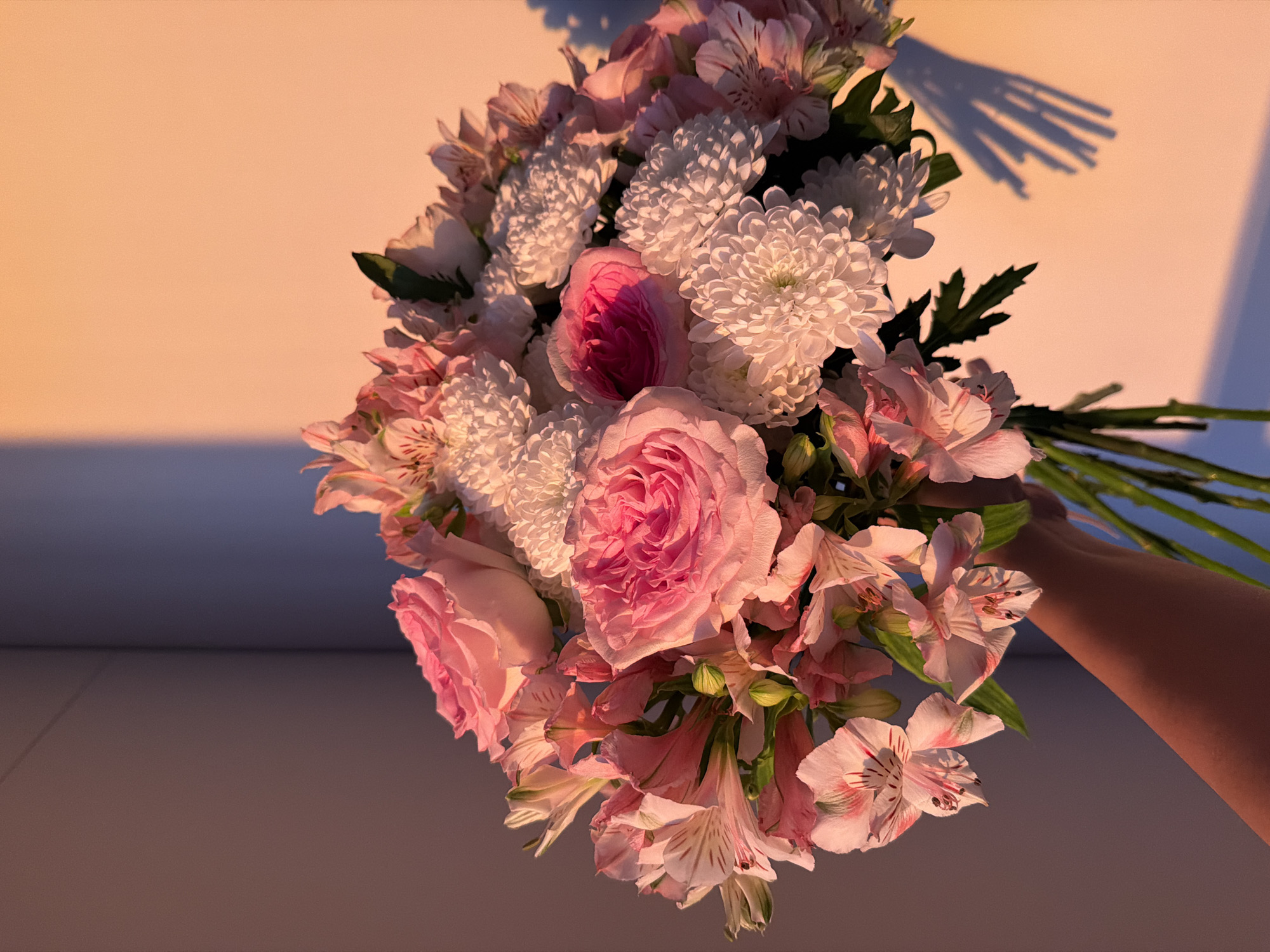 A hand holds a bouquet of pink and white flowers, likely roses, chrysanthemums, and alstroemeria. The background appears to be an interior wall, illuminated with warm light, casting shadows. The setting seems to be indoors. No specific information suggests this scene is in Almaty, Kazakhstan.