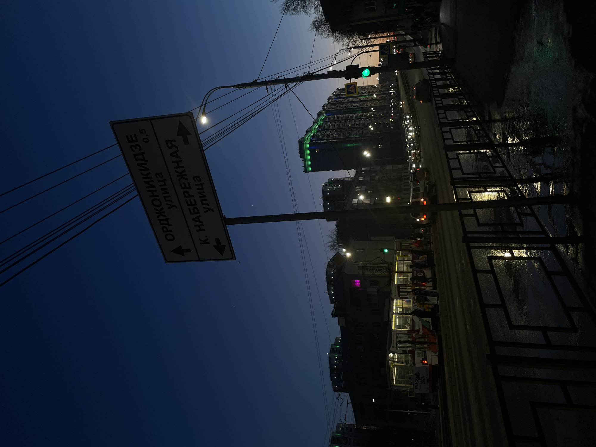 The photograph depicts a nighttime urban scene in Izhevsk, Russia. A street sign indicates directions to "ул. Орджоникидзе" (Ordzhonikidze Street) and "K. Набережная улица" (K. Naberezhnaya Street). A tall apartment building with green illuminated details is visible in the background. A tram or bus with illuminated windows is stopped along the street. Reflections suggest the ground is wet, possibly from rain or snow. A traffic light is showing green. The lighting is dim, with streetlights and building lights illuminating the scene. A fence or barrier is visible in the foreground.
