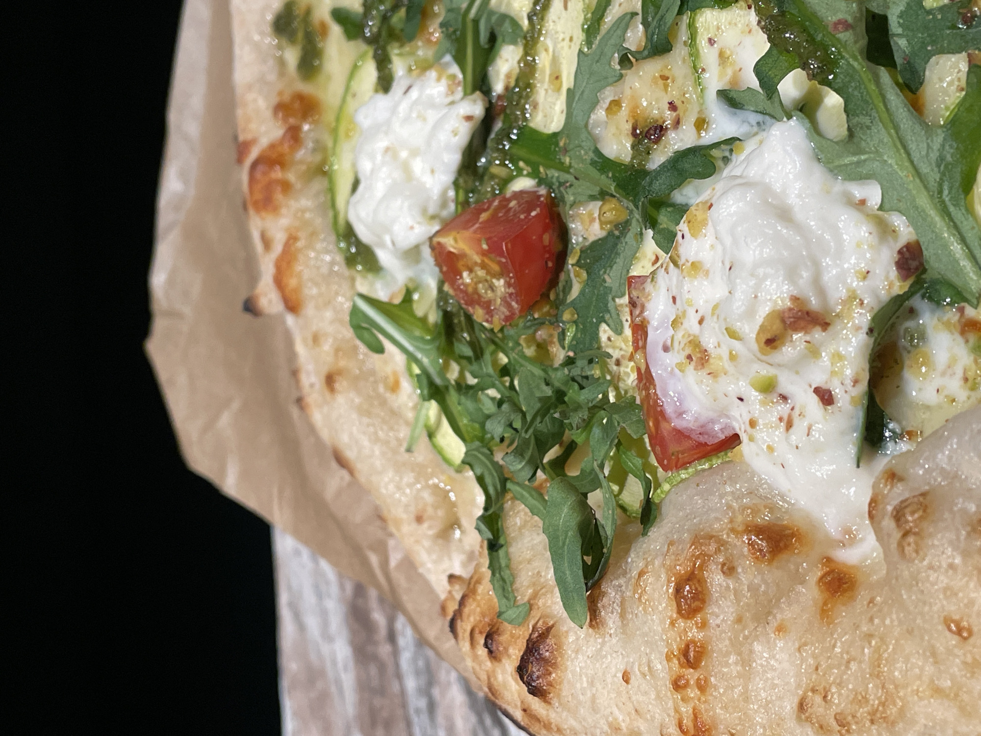 A close-up shot of a pizza on brown parchment paper, possibly in Chaikovsky, Russia. The pizza has a golden crust with browned spots. Toppings include white creamy cheese, green arugula leaves, sliced zucchini, and diced red tomatoes. A green sauce is drizzled over the toppings. The background is black.