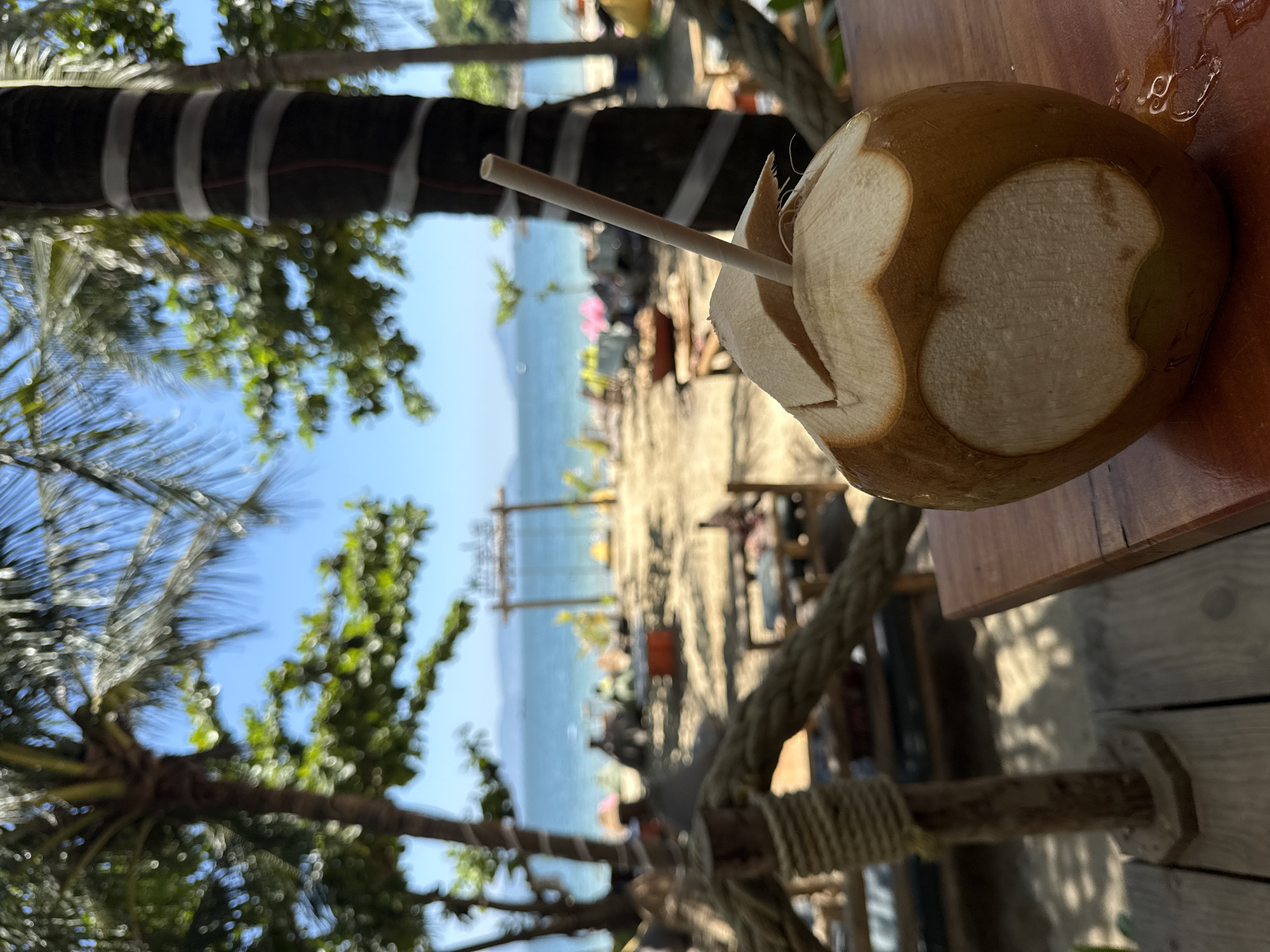 A landscape-oriented photograph shows a partially-opened, young coconut on a wooden table with a brown straw inserted, against a beach backdrop. The coconut, primarily light brown, has a circular opening with a lighter-colored interior. A clear, viscous liquid (potentially coconut water) is visible near the top of the coconut. The straw is made of paper and is a light brown color.
The beach scene includes sand, water, and vegetation. A structure resembling a gate or archway is present, alongside what appear to be beach chairs and people in the distance. The sky is blue with some wispy clouds. The photograph is taken from a slightly elevated perspective. Based on the location, this photograph was taken in Đà Nẵng, Vietnam.