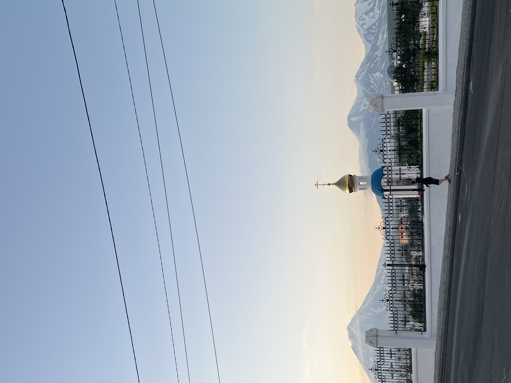 The image shows a street scene in Petropavlovsk-Kamchatsky, Russia, with a view of snow-capped volcanoes in the background. In the foreground, a person is walking along a sidewalk next to a white wall and an ornate black metal fence. Behind the fence, a blue-domed church with a golden cross is visible, along with trees and other buildings. The sky is a clear blue, with power lines strung across the top of the frame. The overall impression is of a peaceful, picturesque landscape.