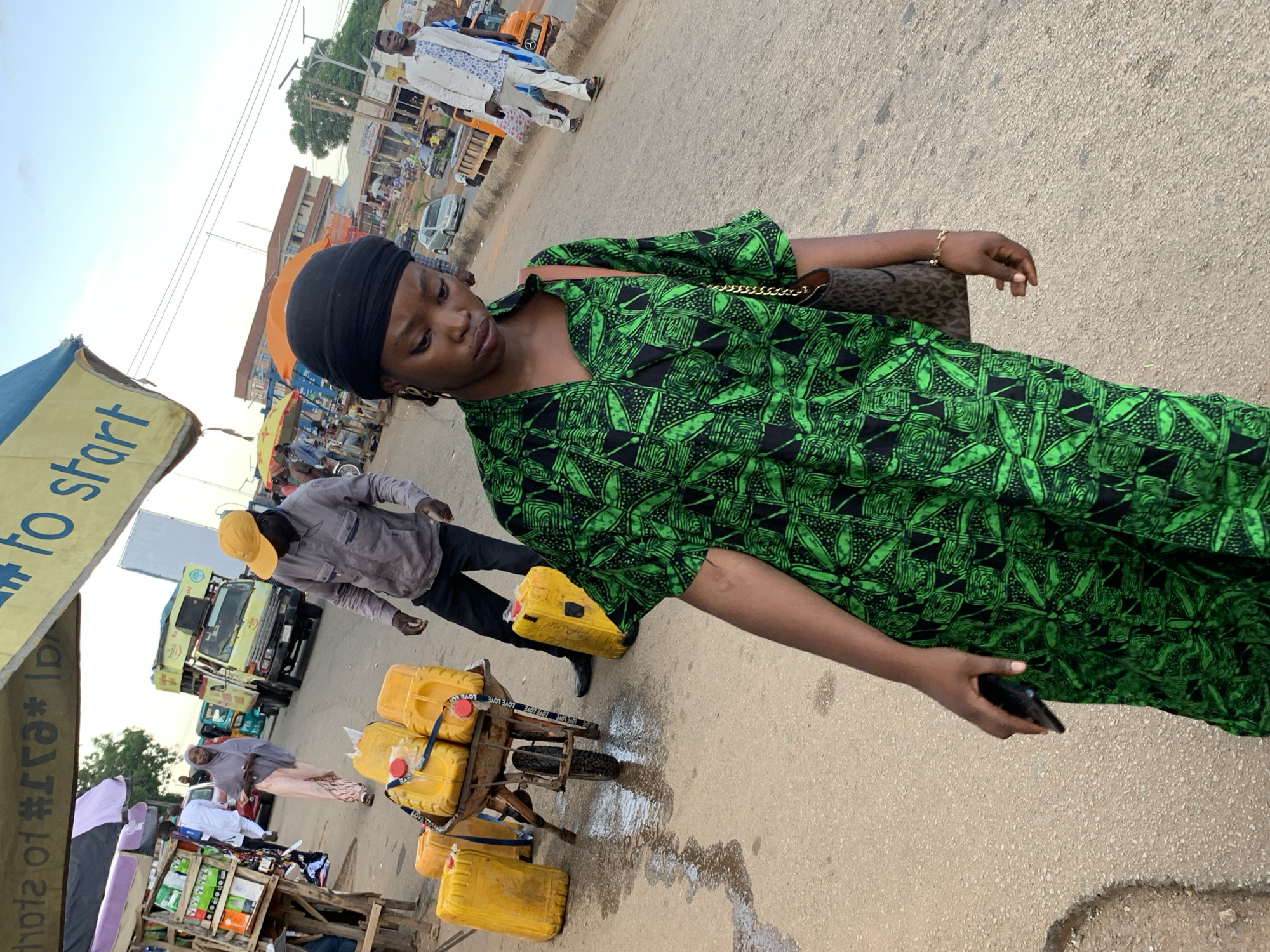 The image depicts a street scene in Jos, Nigeria, with a young woman in the foreground wearing a green and black patterned dress and a black headscarf. She is looking down and holding a smartphone in her left hand. Behind her, a man in a grey shirt and yellow cap is walking away, and a wheelbarrow laden with yellow jerry cans of liquid is visible. In the background, various vehicles, including a truck and a car, are present, along with market stalls and buildings. The ground is paved and appears wet in places. The overall atmosphere suggests a bustling marketplace or street.