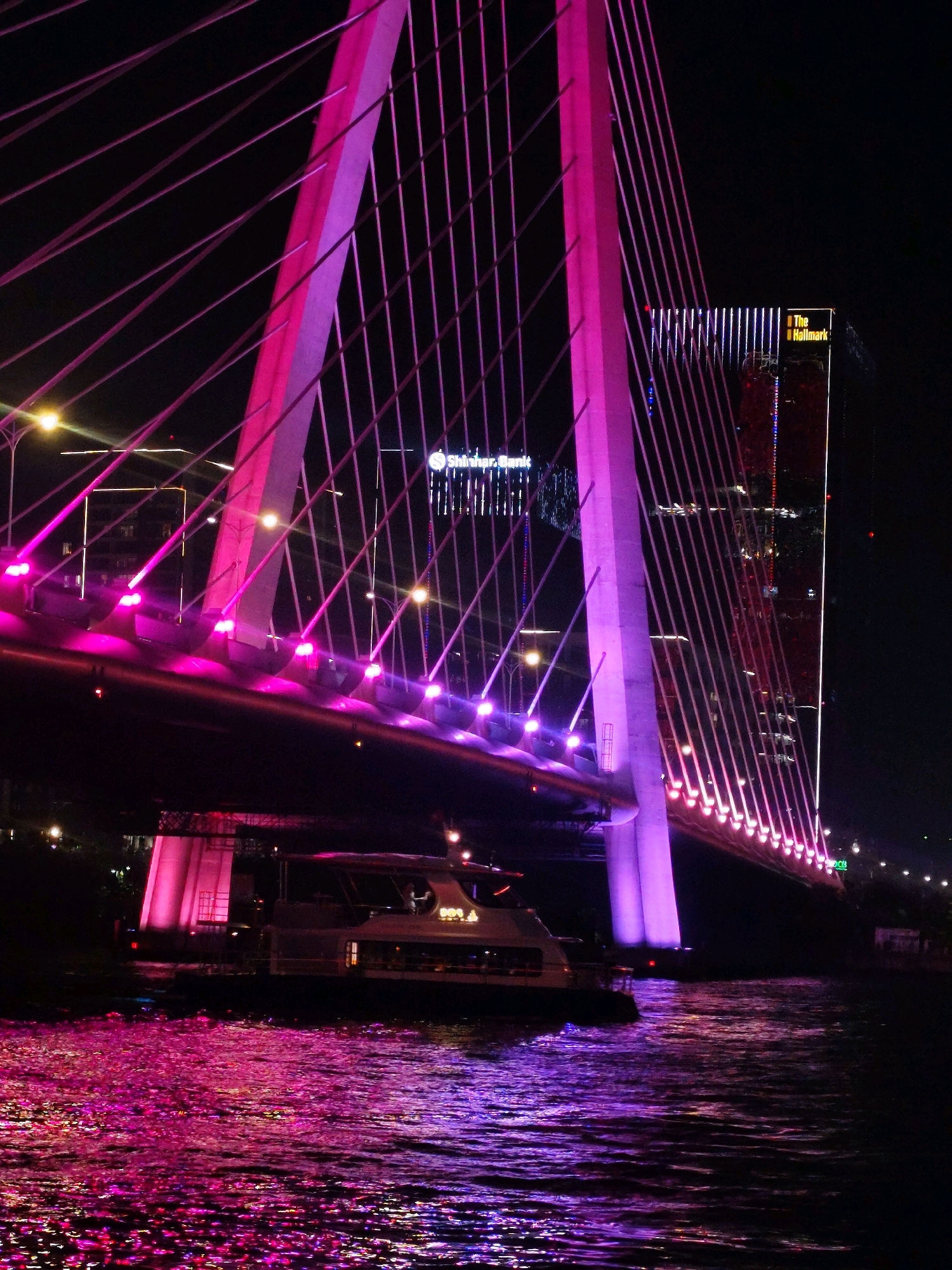 This image captures the Dragon Bridge in Da Nang, Vietnam, illuminated at night with vibrant pink and purple lights. The bridge's distinctive architecture, featuring a dragon-shaped body and a single pylon supporting suspension cables, is prominently displayed. The cables themselves are also lit with pink lights, creating a dazzling effect against the dark sky. In the foreground, the Han River flows, reflecting the colorful illumination from the bridge and the city lights. A white boat with passengers is visible on the water, adding a sense of scale and activity to the scene. In the background, the silhouette of a tall building, likely a hotel or office, is outlined with vertical lines of light. The overall atmosphere is one of modern urban vibrancy and architectural beauty.