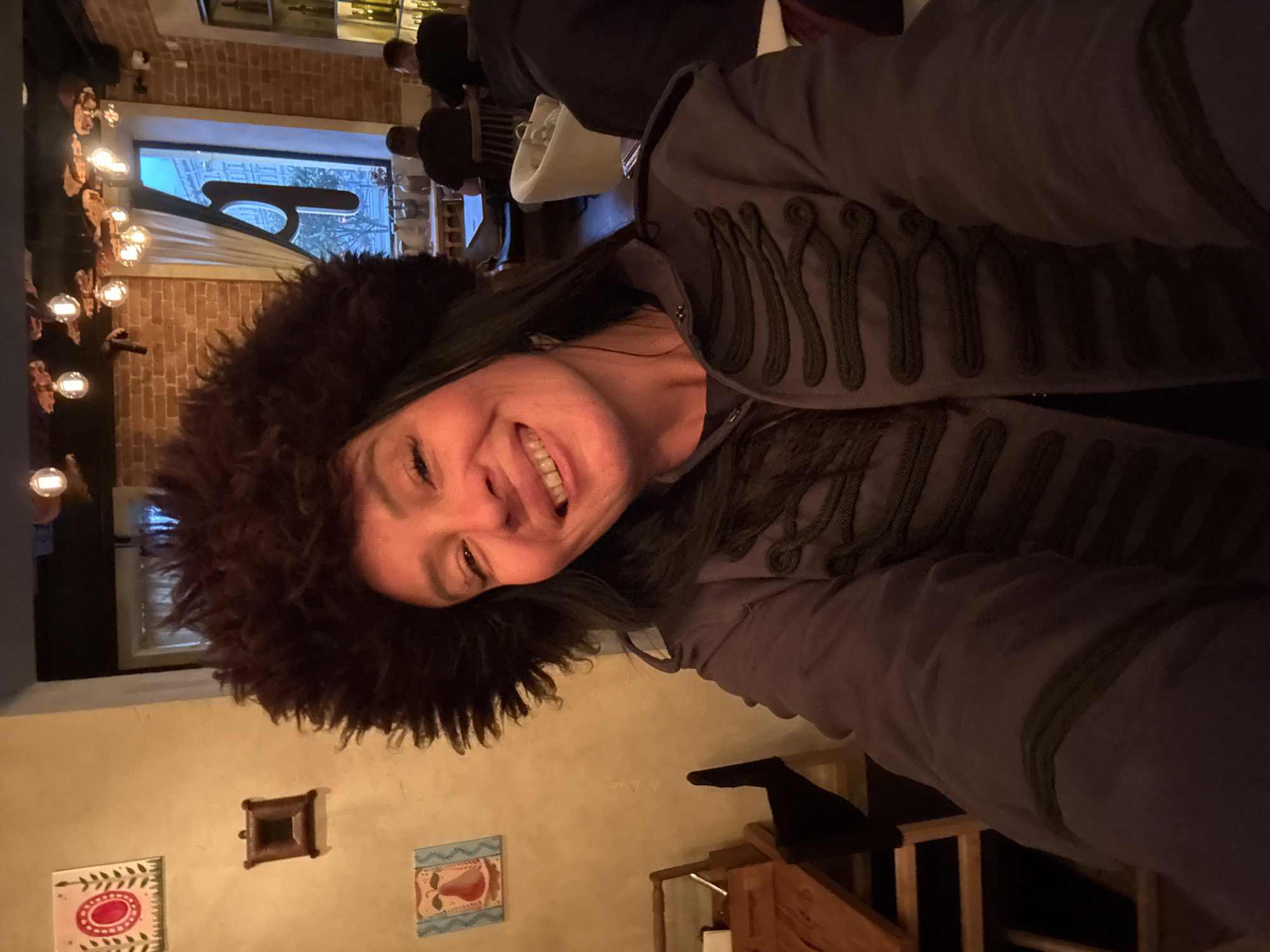 A woman with dark hair, wearing a large, fluffy brown hat and a dark jacket with ornate braiding, smiles at the camera. She is indoors, in what appears to be a restaurant or bar. The background shows exposed brick walls, decorative lighting fixtures, and a window with a view of a city street. There are other patrons in the background, seated at tables. The image has a warm, intimate feel.