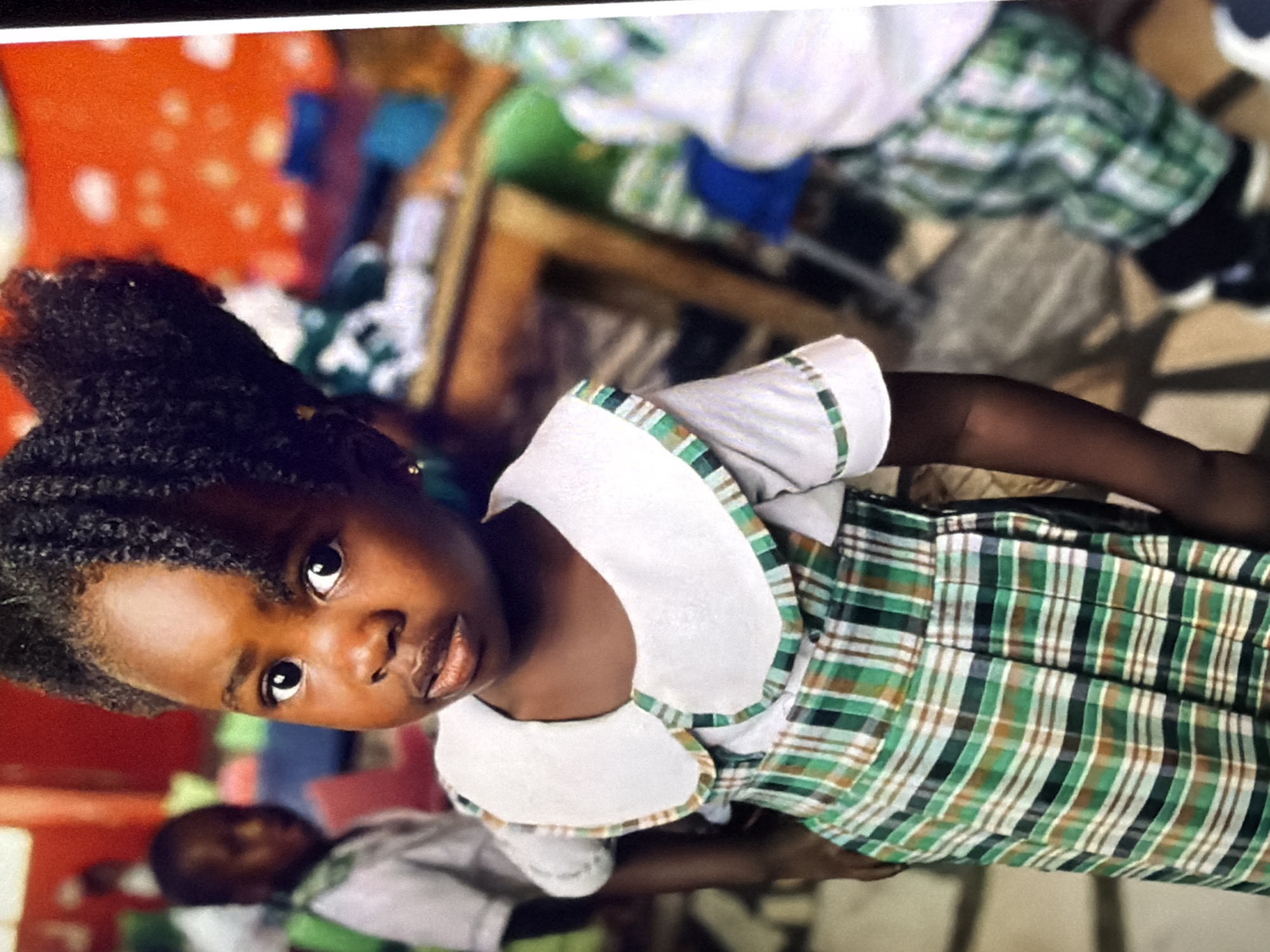 The image is a medium shot of a young Black girl in a classroom setting. She is wearing a white shirt with a large, white collar that has a green and brown plaid trim. Her dress is also made of the same green and brown plaid fabric. Her dark hair is styled in an updo. Her eyes are wide and looking directly at the camera. The background is blurred but appears to be a classroom with colorful elements, possibly educational toys or decorations. Another child is visible in the background, also wearing a school uniform. The lighting suggests it is daytime, likely indoors. The overall mood of the image is captured by the girl's direct gaze, which could be interpreted as curious or perhaps a bit shy. There is no visible text in the image.