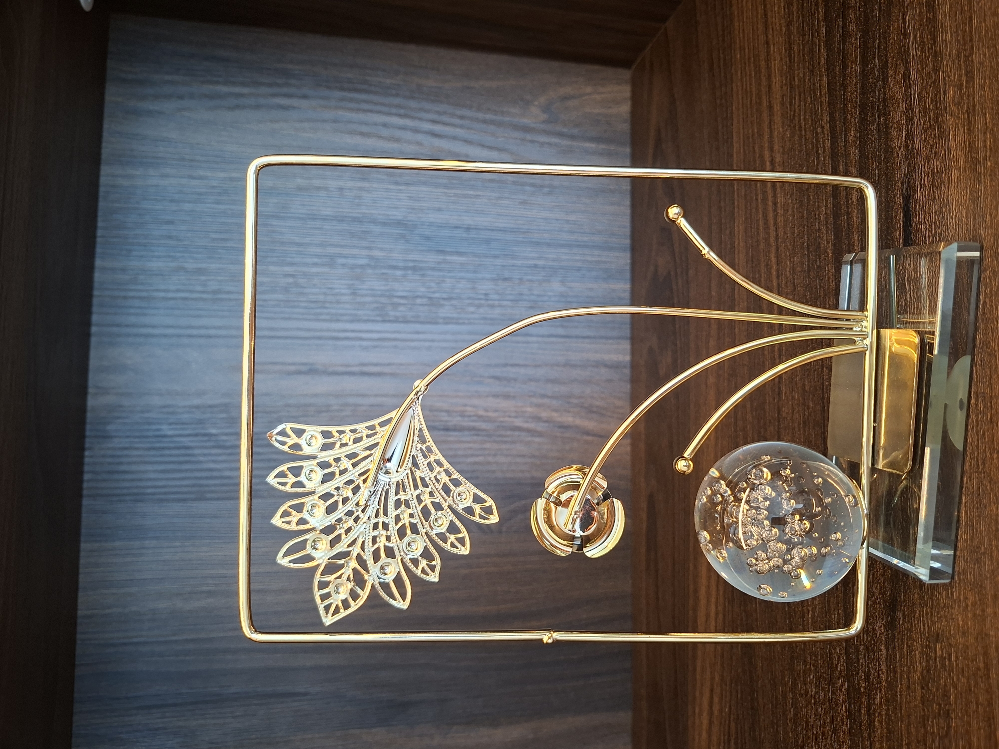 A golden metallic sculpture of a stylized peacock feather is positioned on a dark wooden shelf. The sculpture is framed by a thin, rectangular golden frame. Below the feather, there is a clear glass sphere filled with small bubbles, and to its right, a small golden ball adorns a thin metallic stem. The base of the sculpture is made of clear glass with a golden rectangular piece on top. The background consists of a dark wooden paneled wall. The scene is lit from the front, casting shadows. The object appears to be an ornament or award. The location of West Jakarta, Indonesia is not evident from the image itself, and therefore cannot be included in the description.