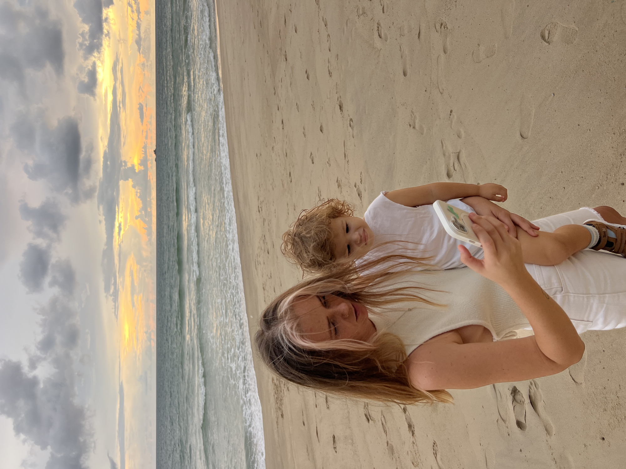 A young woman with long blonde hair, streaked with lighter blonde and pink highlights, is holding a toddler with curly blonde hair. Both are wearing white clothing. The woman is looking down at a smartphone she is holding in her right hand. The toddler is looking away from the camera with a slightly displeased expression. They are standing on a sandy beach with footprints visible. In the background, the ocean meets the sky at sunset. The sky is filled with dramatic clouds in shades of gray, yellow, and orange. A small island or structure can be seen on the horizon in the distance. The overall scene appears to be a serene moment on a beach during dusk.