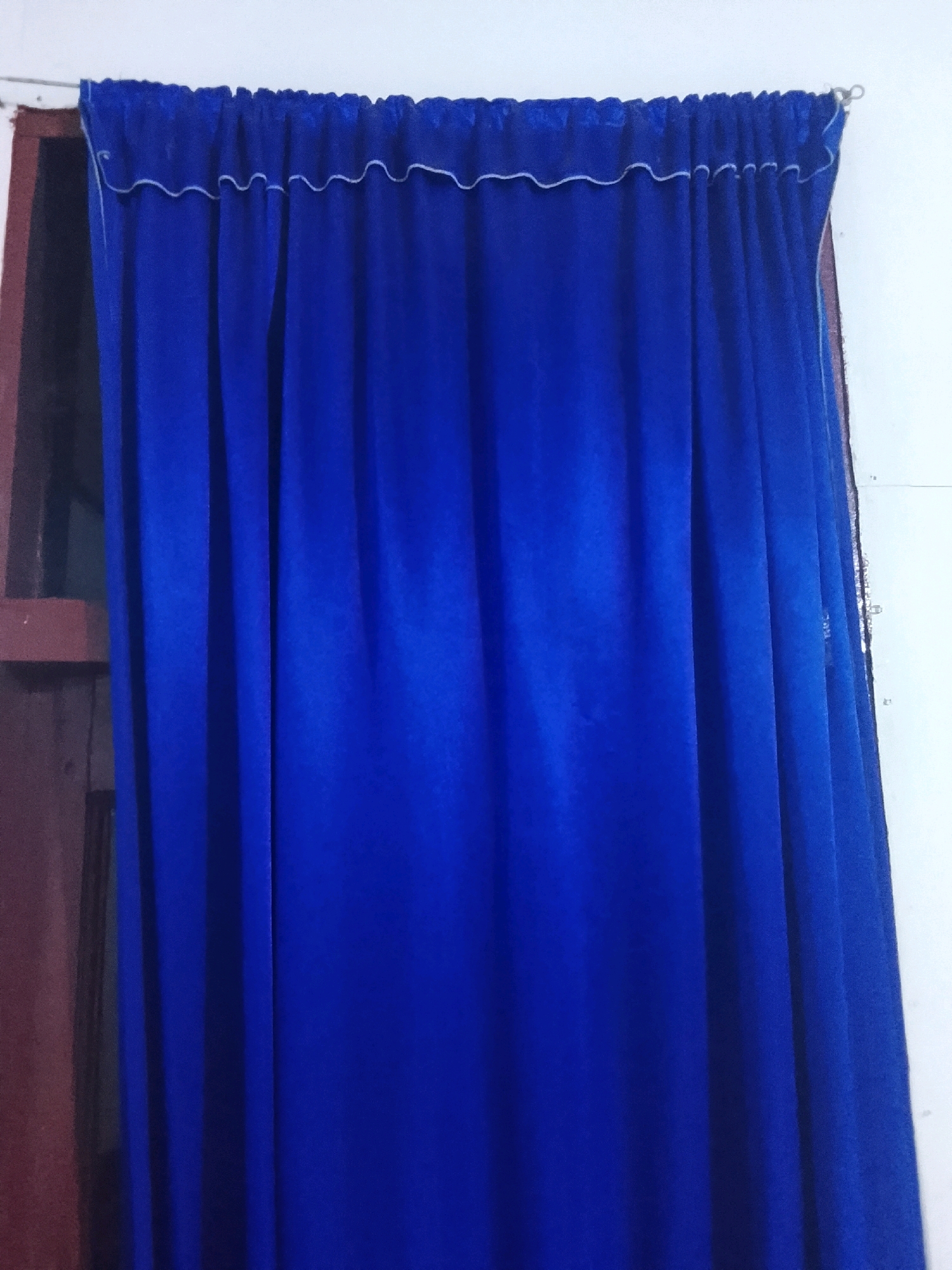 A royal blue curtain with a decorative white trim at the top hangs against a white wall. To the left, a portion of a dark wooden window frame is visible. The curtain is gathered at the top, creating a ruffled effect. The lighting in the image is somewhat dim, casting subtle shadows on the folds of the curtain. The overall impression is of a simple window covering in a domestic setting. The image does not provide any specific information about the city of Lhokseumawe, Indonesia, or any people, actions, or interactions.