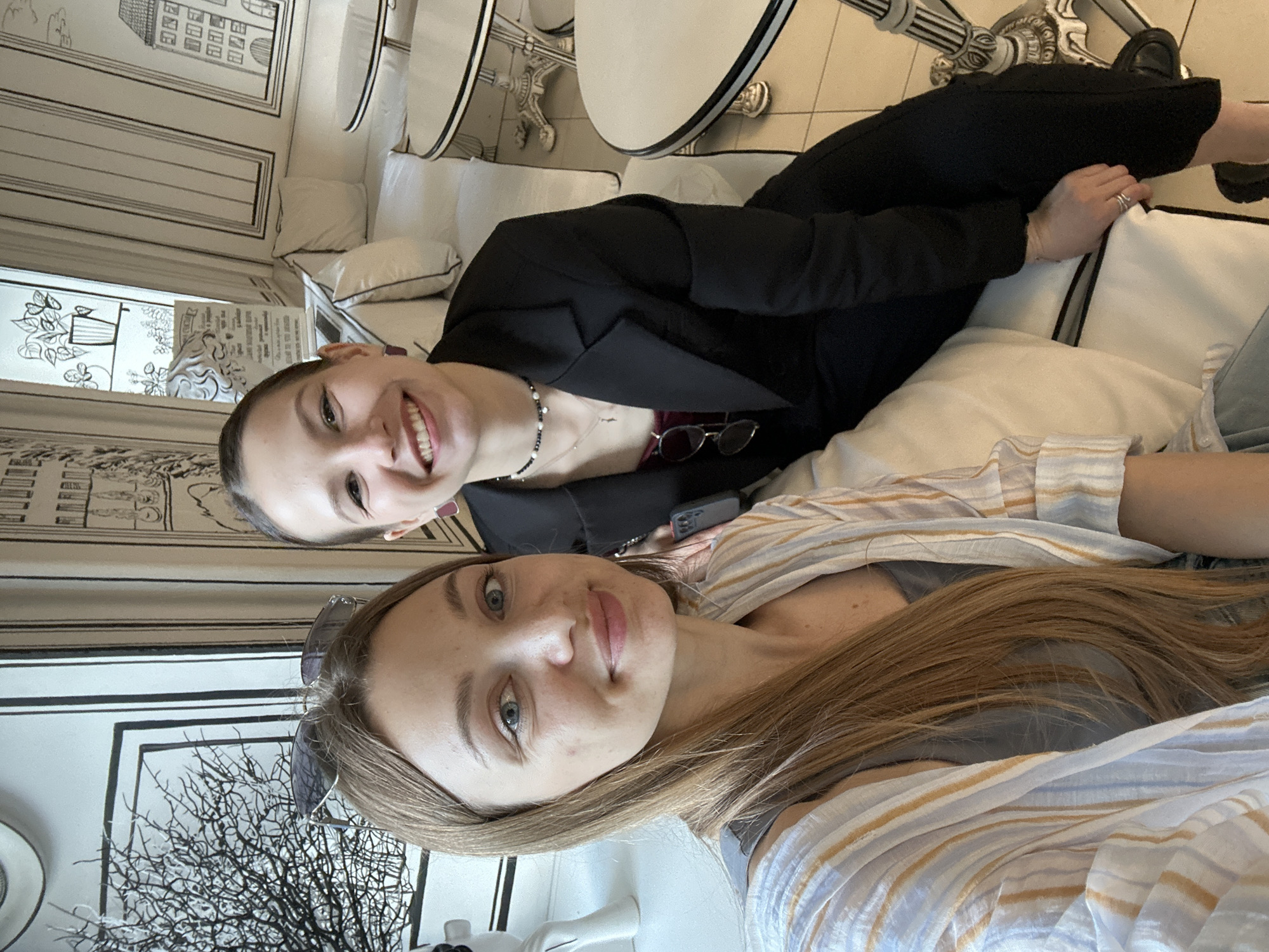 Two women are taking a selfie in a cafe. The woman in the foreground is on the left, with long brown hair and blue eyes, wearing a gray top and a striped cardigan. She is smiling at the camera. The woman in the background is on the right, with dark hair pulled back and blue eyes, wearing a black blazer and pants. She is also smiling at the camera. The cafe has a distinctive black and white cartoon-style decor, with drawings of buildings and plants on the walls. There are white upholstered benches and round tables with white tops. The scene appears to be in Minsk, Belarus, based on visual cues from the cafe's unique design, which is reminiscent of the "2D Cafe" concept that has been observed in the city.