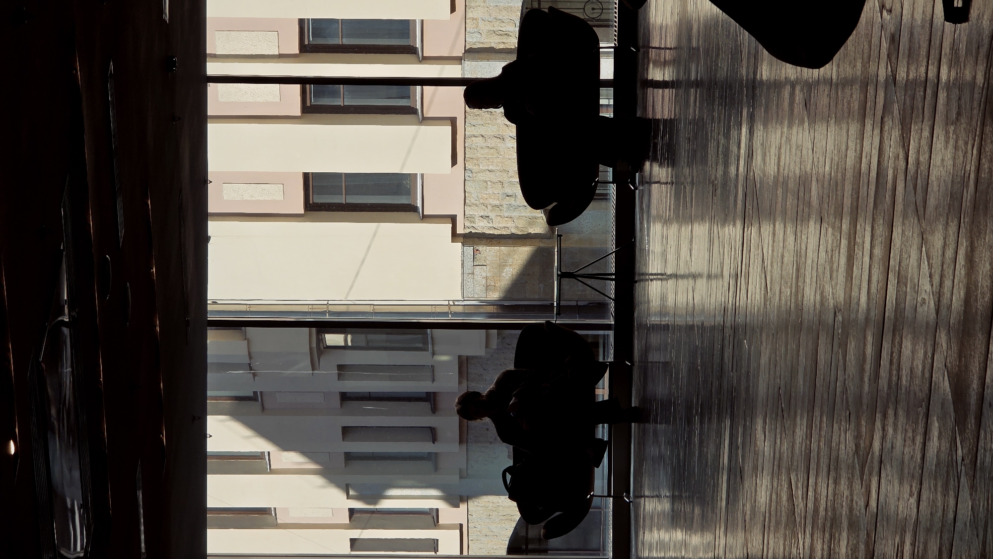 The image depicts an indoor scene looking out through a large window onto an urban environment. In the foreground, two silhouetted figures are seated on high chairs or stools at a counter, facing outwards towards the window. Their features are indistinguishable due to strong backlighting. The foreground also features a reflective surface, likely a wooden floor or a polished countertop, displaying prominent vertical lines and reflections of the exterior.
Through the window, several buildings are visible. One prominent building has a cream-colored facade with rectangular windows framed in a muted reddish-brown hue. Adjacent to it, a section of another building exhibits a textured, light-colored stone or brick cladding. Direct sunlight illuminates parts of these exterior buildings, casting clear shadows, suggesting a bright daytime setting. To the far left of the frame, a dark, vertical element, possibly a door frame or wall panel, is partially visible.
The scene conveys a quiet moment of observation or rest, possibly in a cafe, restaurant, or a public waiting area. The architectural style of the buildings seen through the window is consistent with an urban European setting. There is no visible text in the image.
