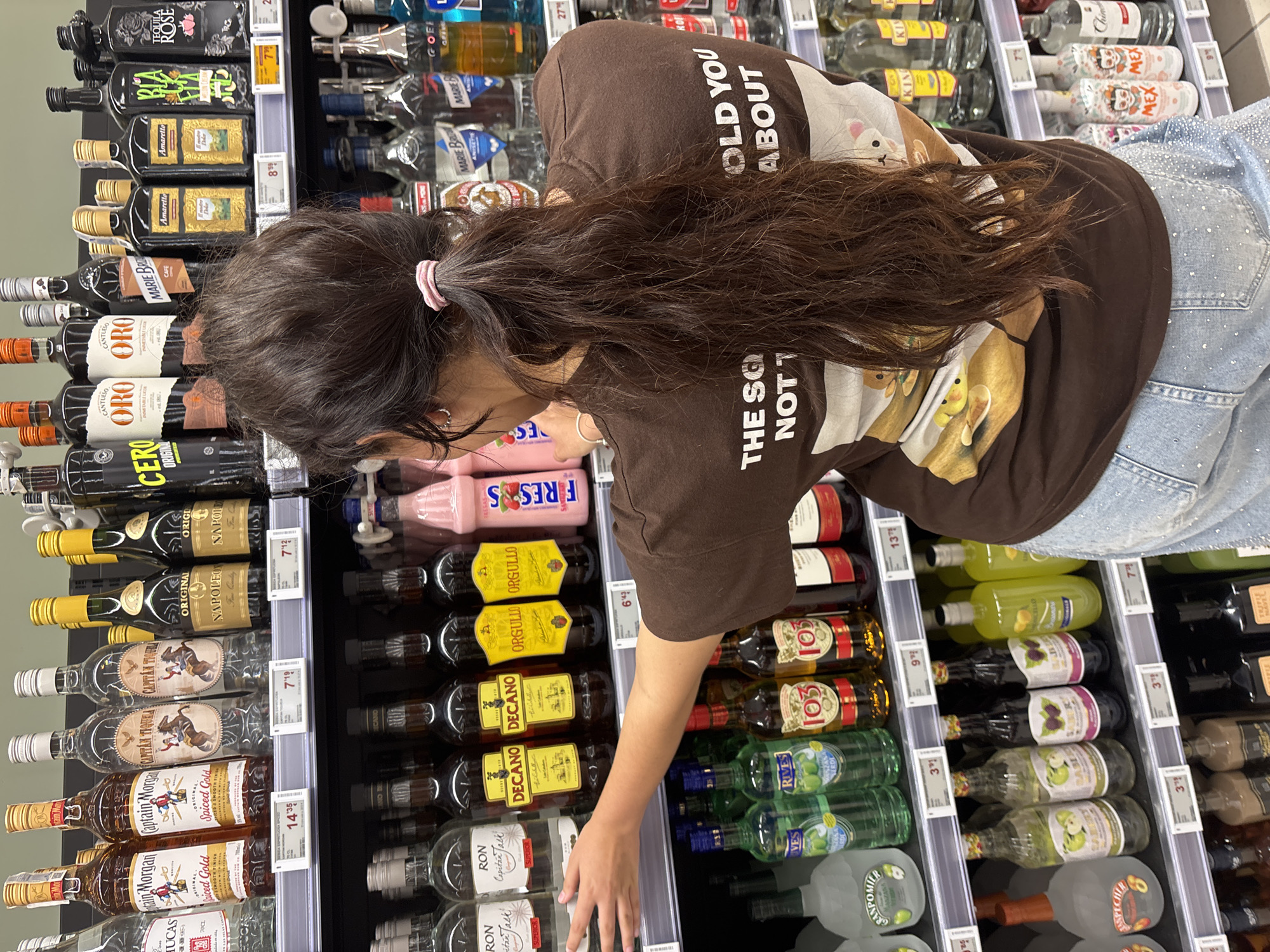 A person with long, dark brown hair, tied in a ponytail with a pink scrunchie, is standing in front of shelves stocked with various bottles of alcoholic beverages in a supermarket. The person is wearing a brown t-shirt with a graphic print on the back that reads "THE SO OLD YOU NOT T ABOUT" and has images of cartoon characters. They are also wearing light blue jeans embellished with rhinestones. The person's left arm is extended, reaching towards the shelves.
The shelves are filled with a variety of liquor bottles, including Captain Morgan Spiced Gold, Captain Morgan White, Tequila Rose, Amaretto, and other brands. Some bottles have yellow and gold labels, while others are clear or dark glass. Price tags are visible on the shelves below some of the bottles. The environment appears to be a retail setting, likely a supermarket or liquor store, with a focus on alcoholic beverages.
The specific location is identified as Urbanización Villa Martín, Spain. The lighting suggests an indoor, well-lit environment typical of a commercial establishment. The image captures a moment of shopping or browsing, with the person interacting with the displayed products.
