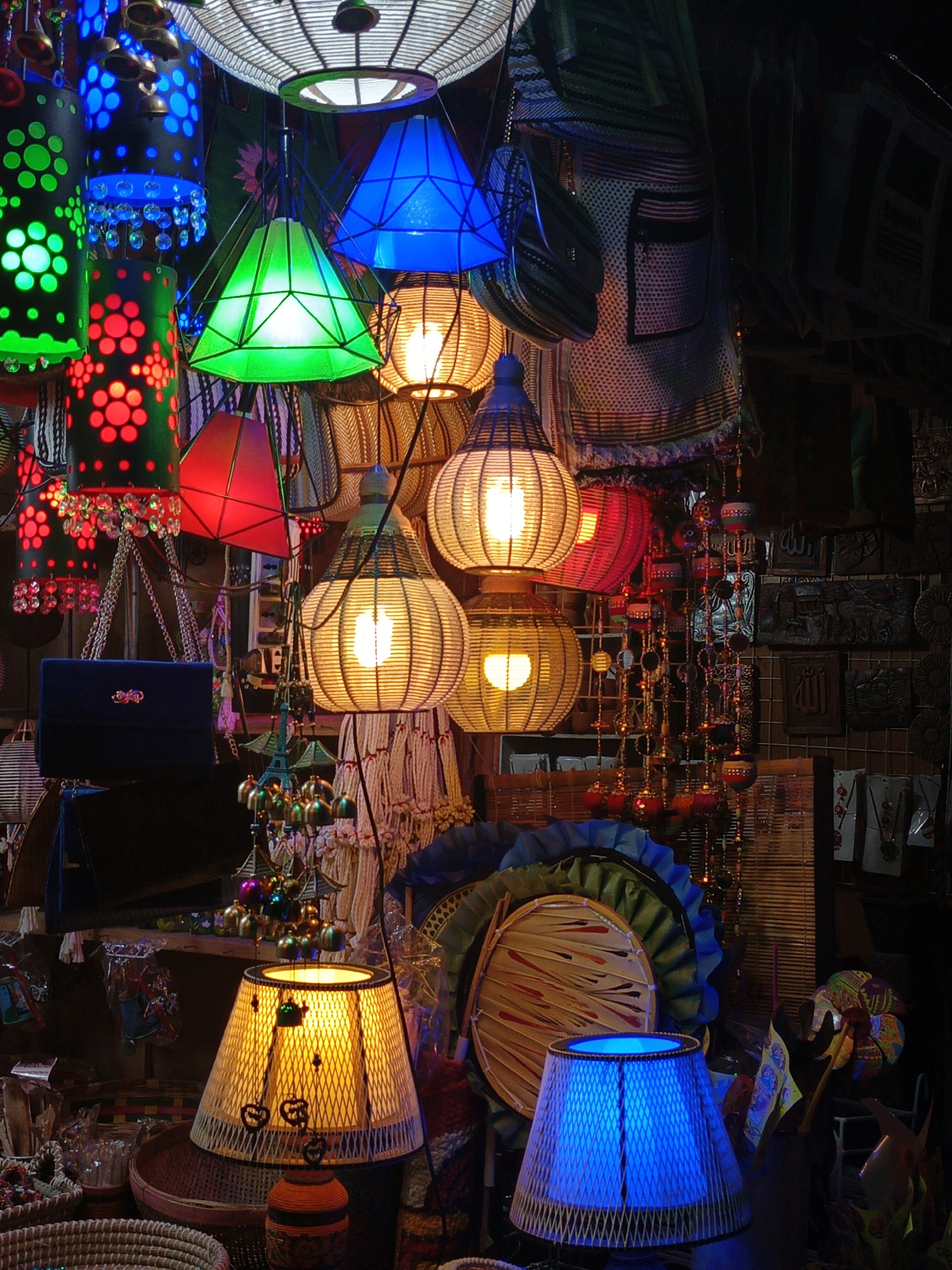 This vibrant photograph captures a richly decorated stall or shop, likely a craft market in Dhaka, Bangladesh, bustling with an array of illuminated lamps and handcrafted goods. The scene is dominated by a diverse collection of lamps, glowing in various colors and designs.
Hanging from the ceiling are numerous lanterns: cylindrical ones in red, green, and blue, featuring intricate perforated patterns and often adorned with dangling crystal-like beads. Geometric pyramid-shaped lamps, in bright green and electric blue, also contribute to the overhead display. Further down, multiple spherical and teardrop-shaped lamps, crafted from woven materials like rattan or wicker, emit a warm, inviting yellow light. Two similar table lamps are visible at the bottom of the frame, one glowing yellow and the other a striking blue.
Interspersed among the lamps are other artisanal items: patterned woven bags, a dark blue velvet clutch bag with a golden embellishment, and strings of colorful decorative bells and beads, some forming ornate wind chimes. In the darker background on the right, several carved wooden panels with what appears to be faint Arabic calligraphy are displayed. Other items include woven baskets and decorative fans.
The prominent artificial lighting suggests the scene is set during evening hours or indoors, creating a warm and lively atmosphere. No people are visible, focusing attention entirely on the intricate and colorful merchandise, indicative of a marketplace showcasing traditional craftsmanship.