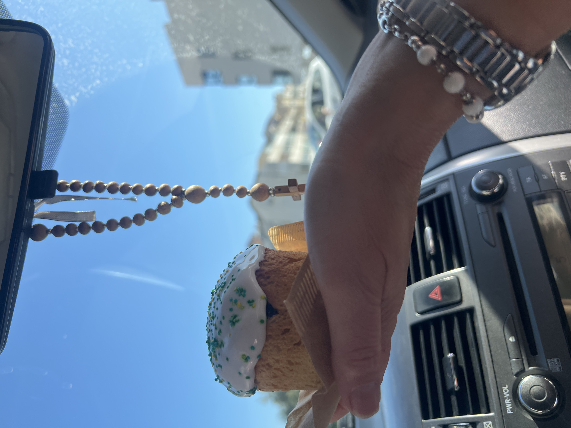 The image depicts an Easter cake held in a hand with a silver bracelet in the interior of a car. A string of wooden beads with a cross hangs from the rearview mirror. In the background, visible through the windshield, is a light blue sky and a building, likely a church, which suggests the location is Budva, Montenegro, considering the cake is often associated with Easter, and the church architecture is reminiscent of the region.