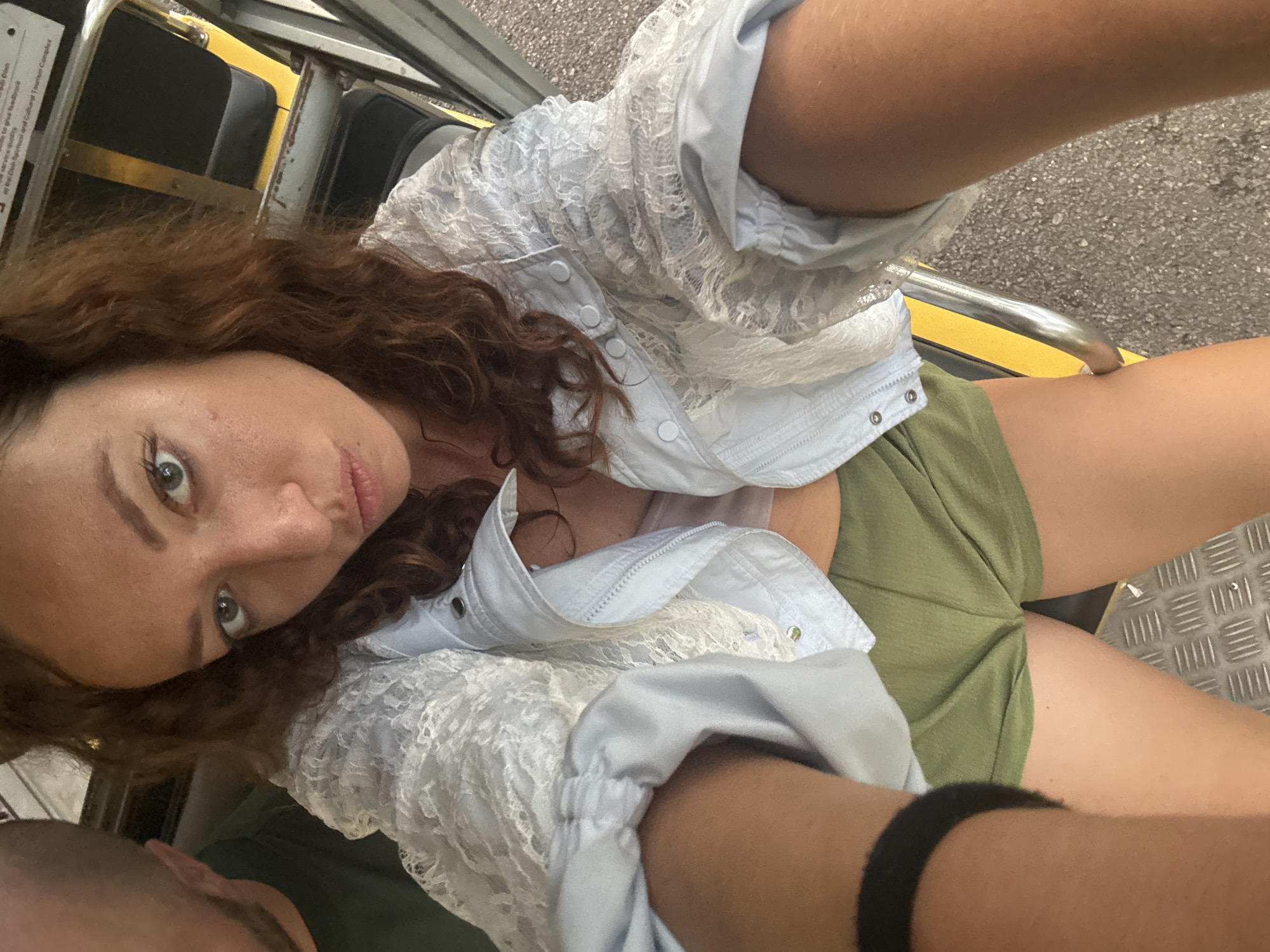 A woman with reddish-brown curly hair is taking a selfie. She is wearing a light blue jacket with white lace detailing and olive green shorts. Her left arm, wearing a black wristband, is visible in the foreground. Another person's head and shoulder, seen from the side, are partially visible to her left. The background appears to be the interior of a vehicle, possibly a bus, with yellow and black striped poles and metal flooring. The image quality is slightly blurry, suggesting it was taken with a mobile device. There is no specific indication of the location within Tây Hoa Lư Ward, Vietnam.