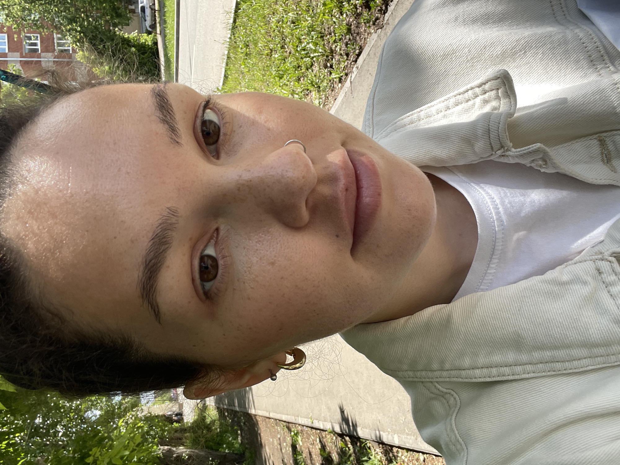 A close-up portrait of a fair-skinned woman with brown hair and eyes, likely in her early 20s, is positioned against an outdoor background. She is wearing a white t-shirt under a light-colored jacket. A silver nose ring is visible, and she has gold hoop earrings in one ear, along with what appears to be a second, smaller earring. The background consists of a sidewalk or path, greenery, and a building in the distance. The image appears to be taken in Izhevsk, Russia, based on the architectural style of the distant building.