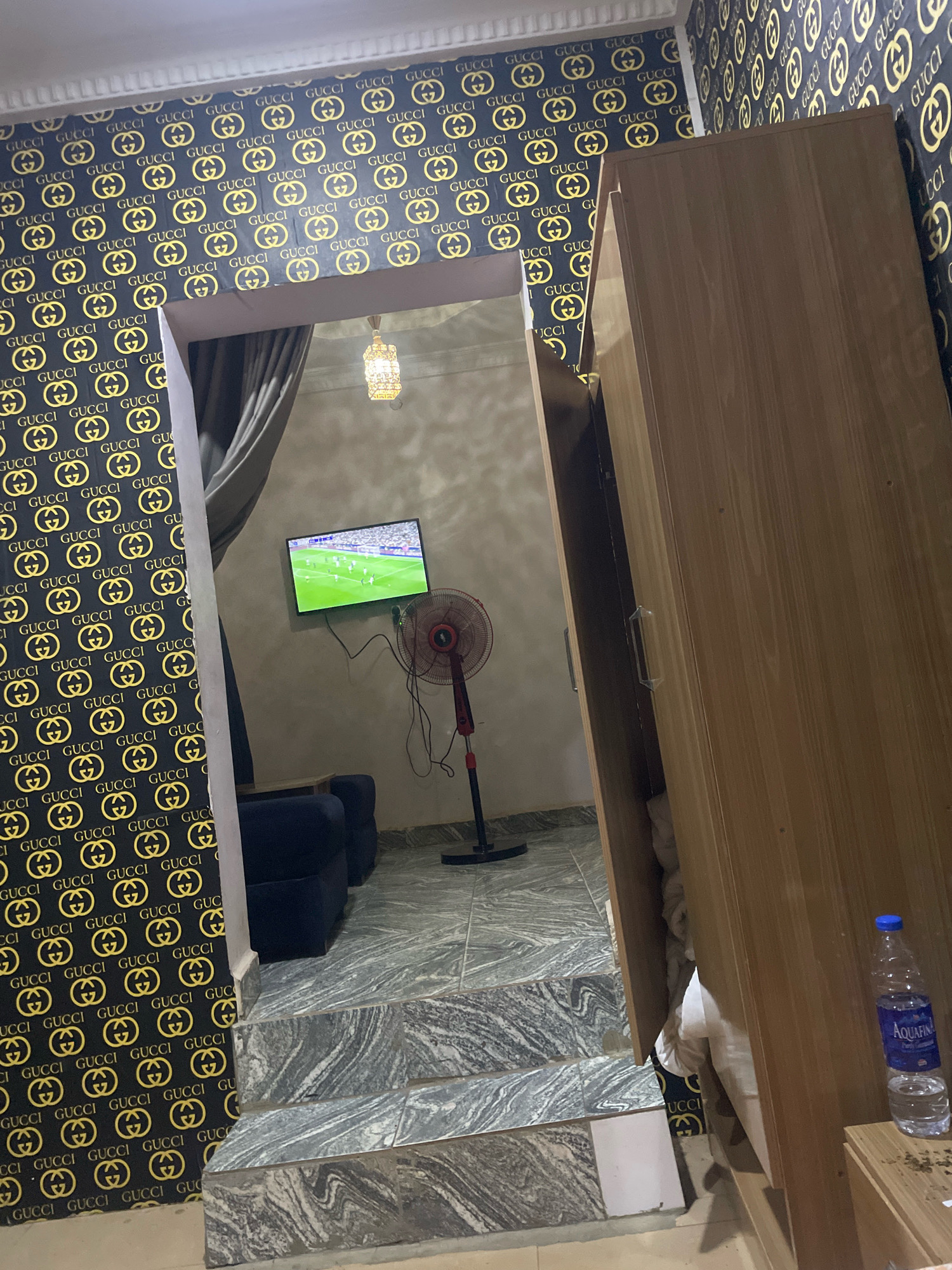 The image depicts an indoor scene, likely a residential room, featuring a prominent Gucci-patterned wallpaper. The room appears to be a hallway or entryway leading into another space. In the foreground, to the right, is a tall, wooden wardrobe, partially open, revealing white bedding and more of the Gucci wallpaper. A bottle of Aquafina water is visible on the wardrobe's surface. To the left, the Gucci wallpaper covers the walls. A reflective surface, likely a mirror or glass partition, is angled, showing a reflection of the space beyond. This reflected space includes a television displaying a soccer game, a red and black standing fan, and a dark blue sofa. A decorative light fixture hangs from the ceiling in the reflected area. The floor is covered in marble-patterned tiles, with a set of marble-patterned steps visible in the foreground leading up to the reflected space. The overall impression is of a modern, possibly decorative, interior. The location is stated to be in the city of Abuja, Nigeria.