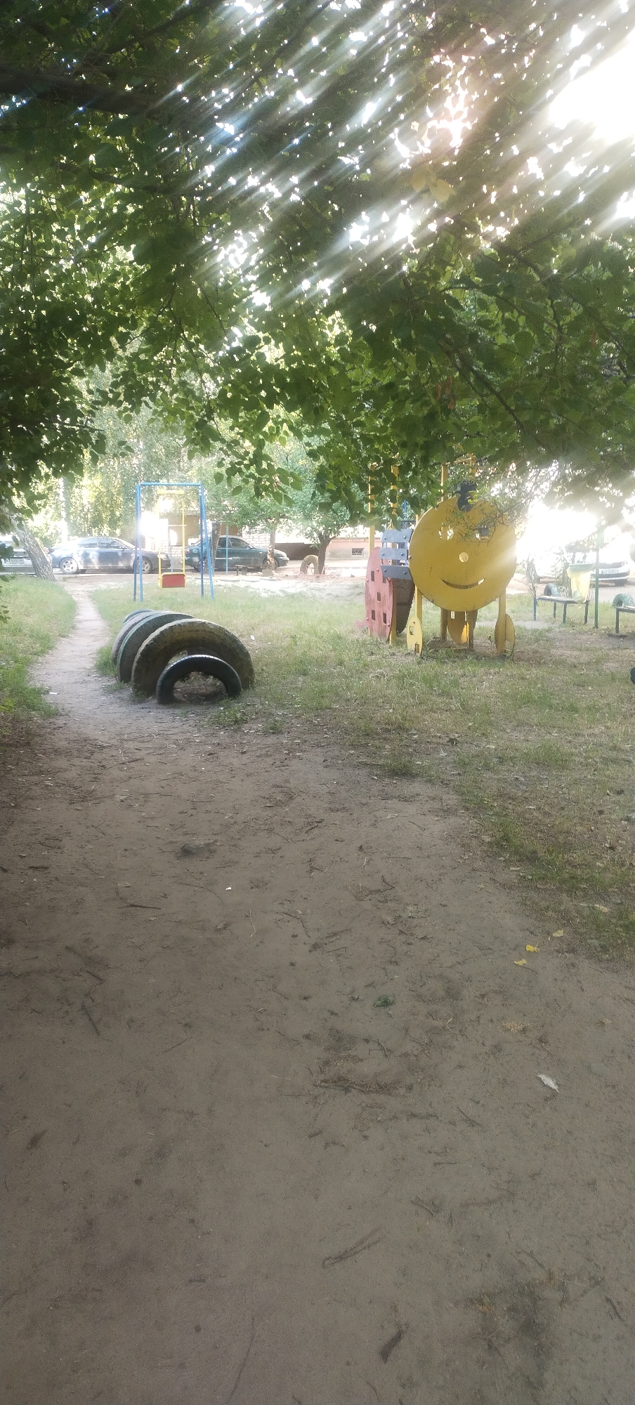 The image captures a playground scene under a canopy of green trees, with sunlight filtering through the leaves. In the foreground, a dirt path is visible, leading towards the playground. Along the left side of the path, three stacked tires serve as a makeshift obstacle or decoration.
Further into the playground, a blue and yellow swing set with a red seat is present. To the right of the swing set, a large, yellow, circular structure with a smiley face design stands prominently. This structure appears to be part of a larger playground apparatus, with some red and pink elements attached to its side. In the background, several parked cars are visible, suggesting the playground is located in a residential area.
The ground of the playground is covered with a mix of grass and dirt. Several benches are scattered throughout the area, some appearing more weathered than others. The overall impression is of a slightly worn but functional outdoor recreational space, likely in a community or apartment complex setting. The presence of cars and the style of the playground equipment suggest an urban or suburban environment. Based on the context provided, this scene is located in the city of Pisochyn, Ukraine.