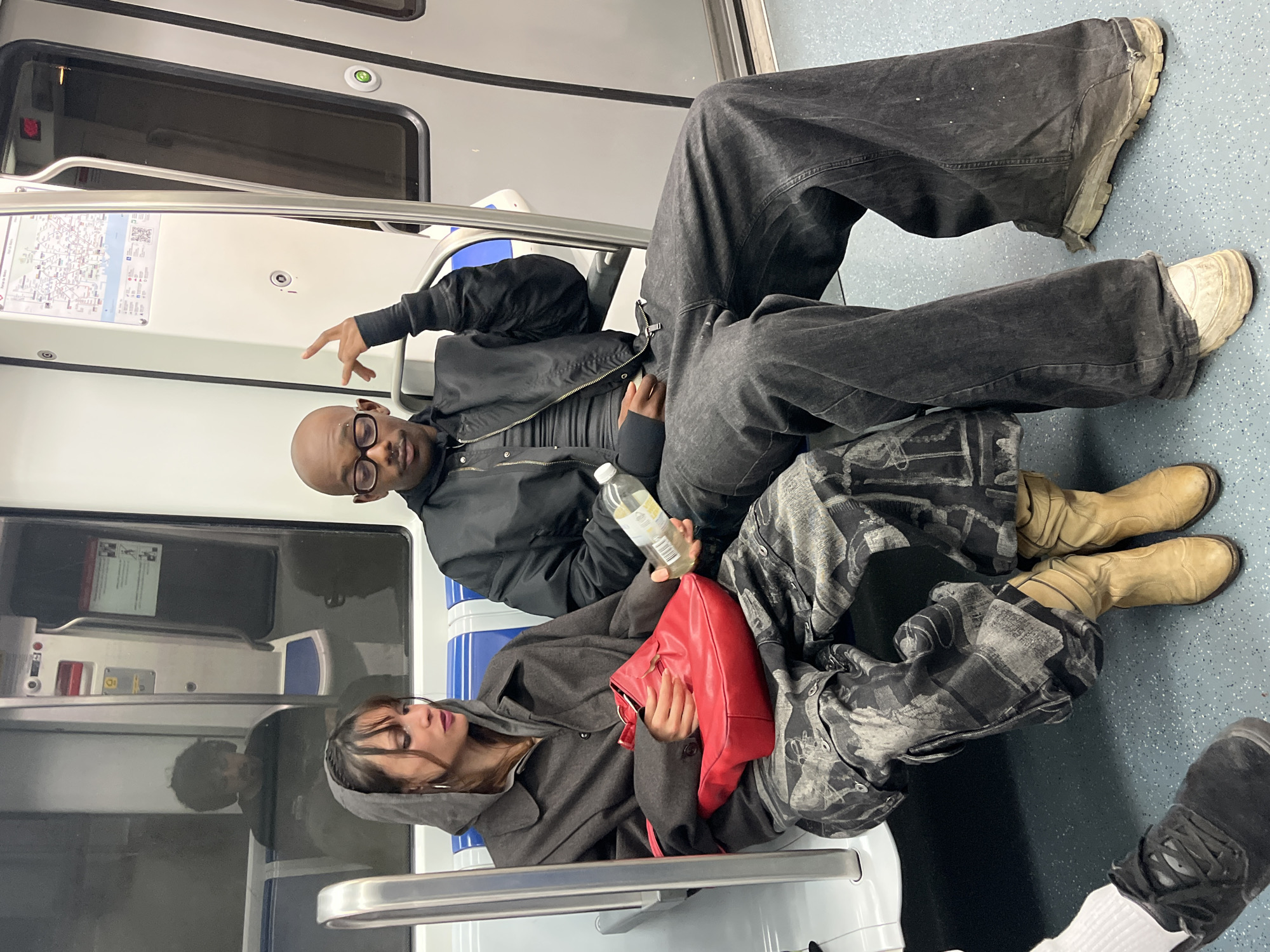 The image shows three people inside a subway car. Two of them are seated and taking up most of the space on one side of the car. The third person's legs and shoes are visible. The person closest to the camera is wearing a hooded gray coat and patterned pants with tan boots, holding a red purse, and appears to be resting. The person next to them is wearing a black jacket, black pants, glasses, and is holding a water bottle. One leg is stretched out towards the camera. The seats are blue and white, and there is a map on the wall of the train. Based on the subway map shown, this is in the city of Barcelona, Spain.