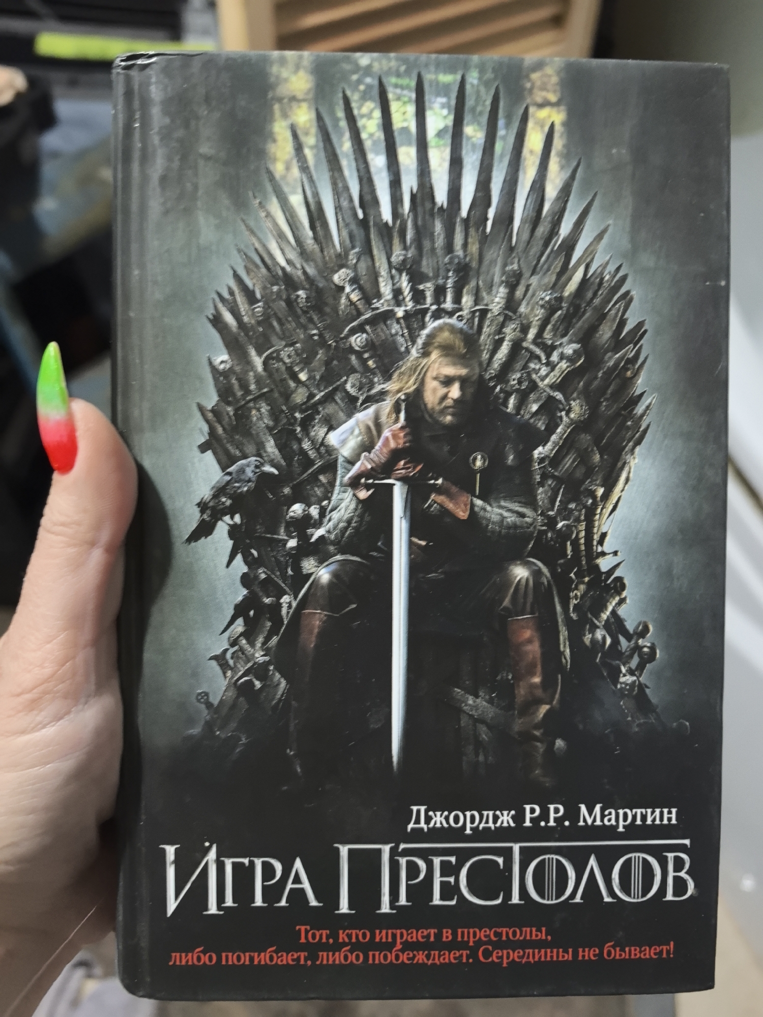 The image shows a hand holding a book. The book is a Russian edition of George R. R. Martin's "A Game of Thrones", titled "Игра Престолов". The cover depicts a man, likely Ned Stark, sitting on the Iron Throne. The throne is made of swords, and a raven is perched on it. The text on the cover includes the author's name and a quote: "Тот, кто играет в престолы, либо погибает, либо побеждает. Середины не бывает!" (He who plays in the thrones, either dies, or wins. There is no middle ground!). The hand holding the book has a nail painted with a red and green gradient. The setting is indoors. There is no information to identify the location as Noviy, Russia.