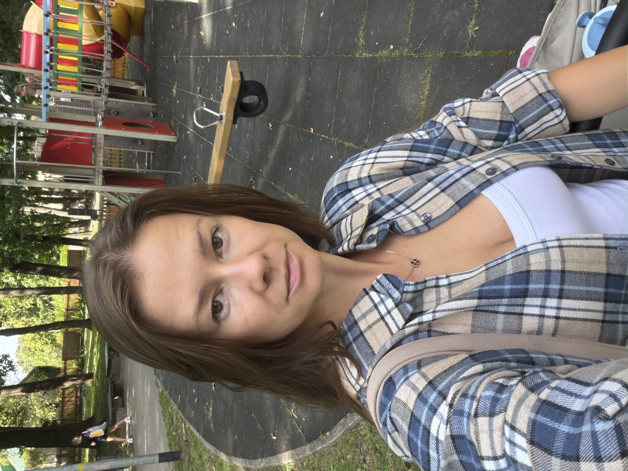In Riga, Latvia, a woman is taking a selfie in a park with a playground. She is wearing a blue and beige plaid shirt over a white tank top and a necklace with a black clover pendant. In the background, a child is walking away from the camera, and part of a colorful playground structure is visible, as well as a seesaw. The ground is a dark, rubberized surface, with a paved pathway and grass also present in the park.