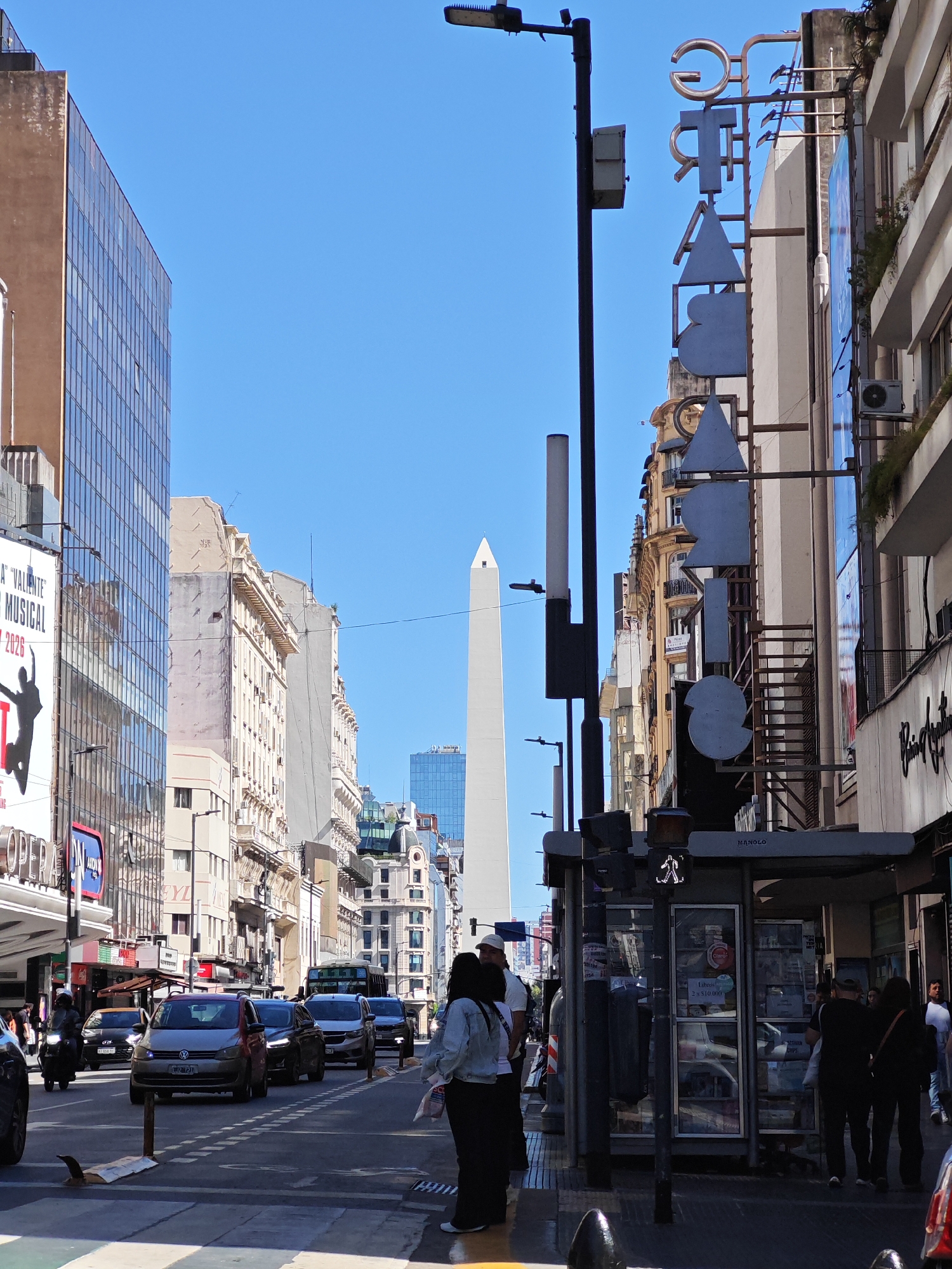 The image depicts a street scene in Buenos Aires, Argentina, with the Obelisco monument visible in the distance. The foreground shows pedestrians on a sidewalk, a newsstand, and traffic on the street. Buildings line both sides of the street, including a building with a large theater sign. The sky is clear and blue. The Obelisco is a tall, white, four-sided obelisk. Cars, including a gray Volkswagen, are moving on the street. A person on a scooter is also visible. The overall impression is of a busy city street on a sunny day.